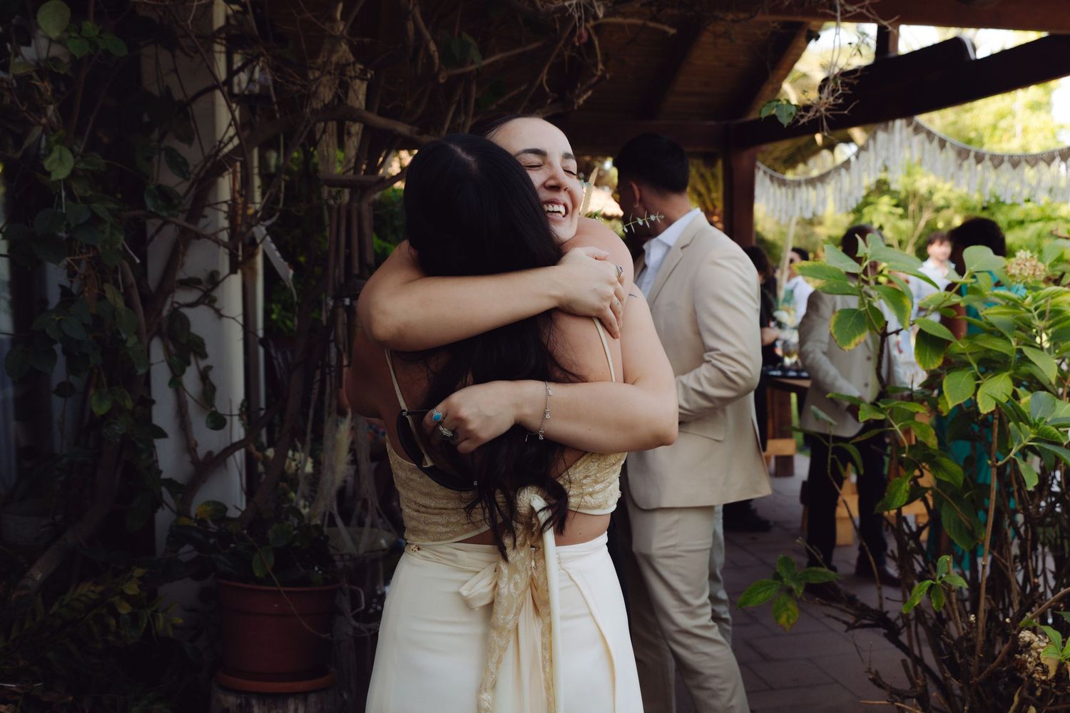 Bride in white dress joyfully hugging a guest at an outdoor wedding reception surrounded by lush greenery.