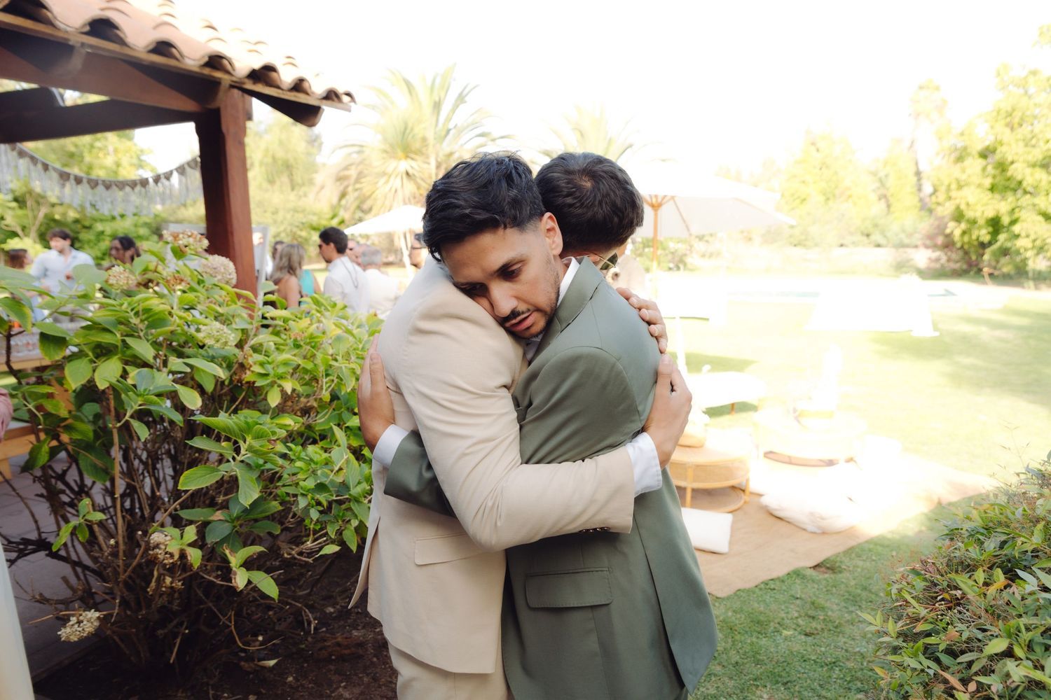 Groom in beige suit sharing a warm embrace with a guest in olive green at an outdoor wedding.
