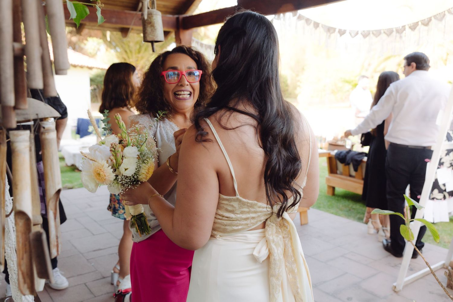 Wedding guest in pink dress smiling and greeting bride in white gown holding a bouquet at outdoor ceremony.