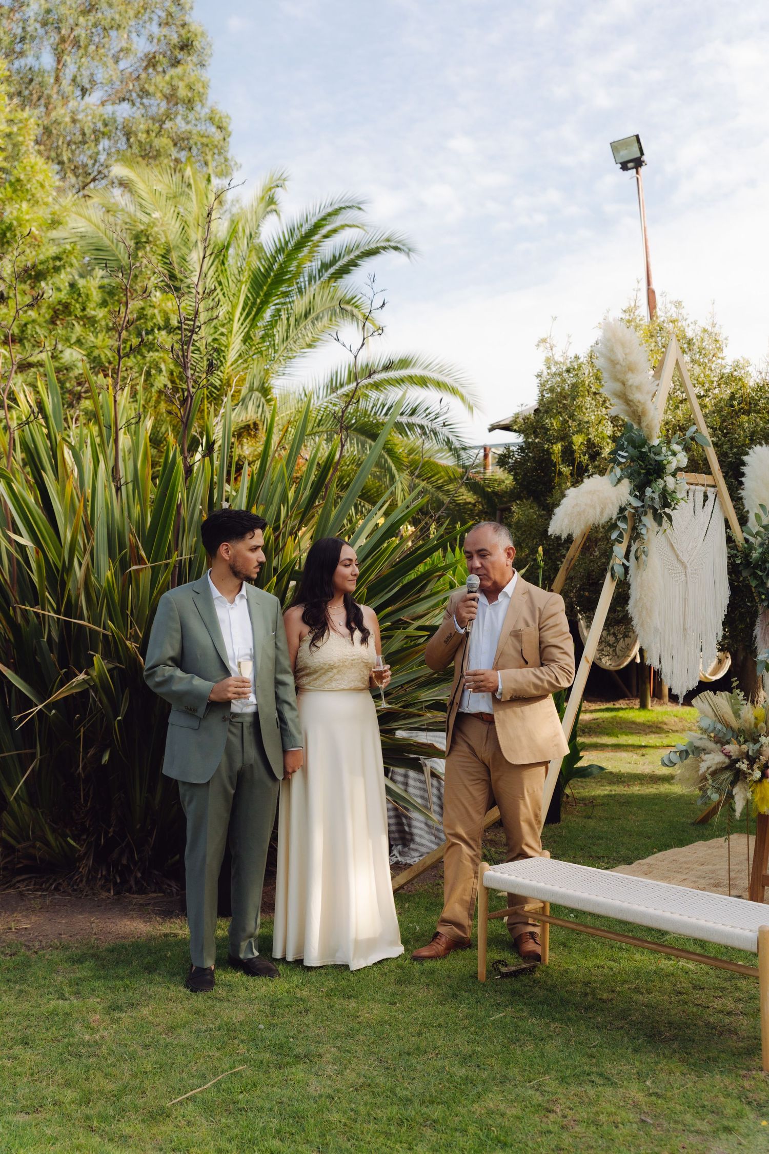 Bride and groom in elegant attire chatting with older guest beside tropical plants at outdoor wedding.