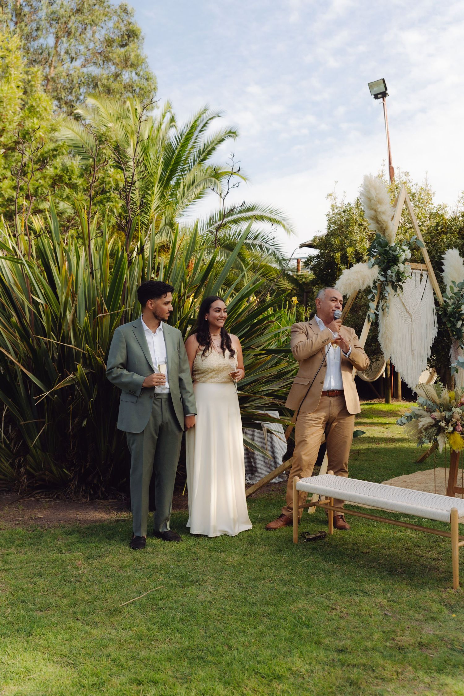 Bride and groom standing with older man giving a speech at a lush outdoor garden wedding reception.