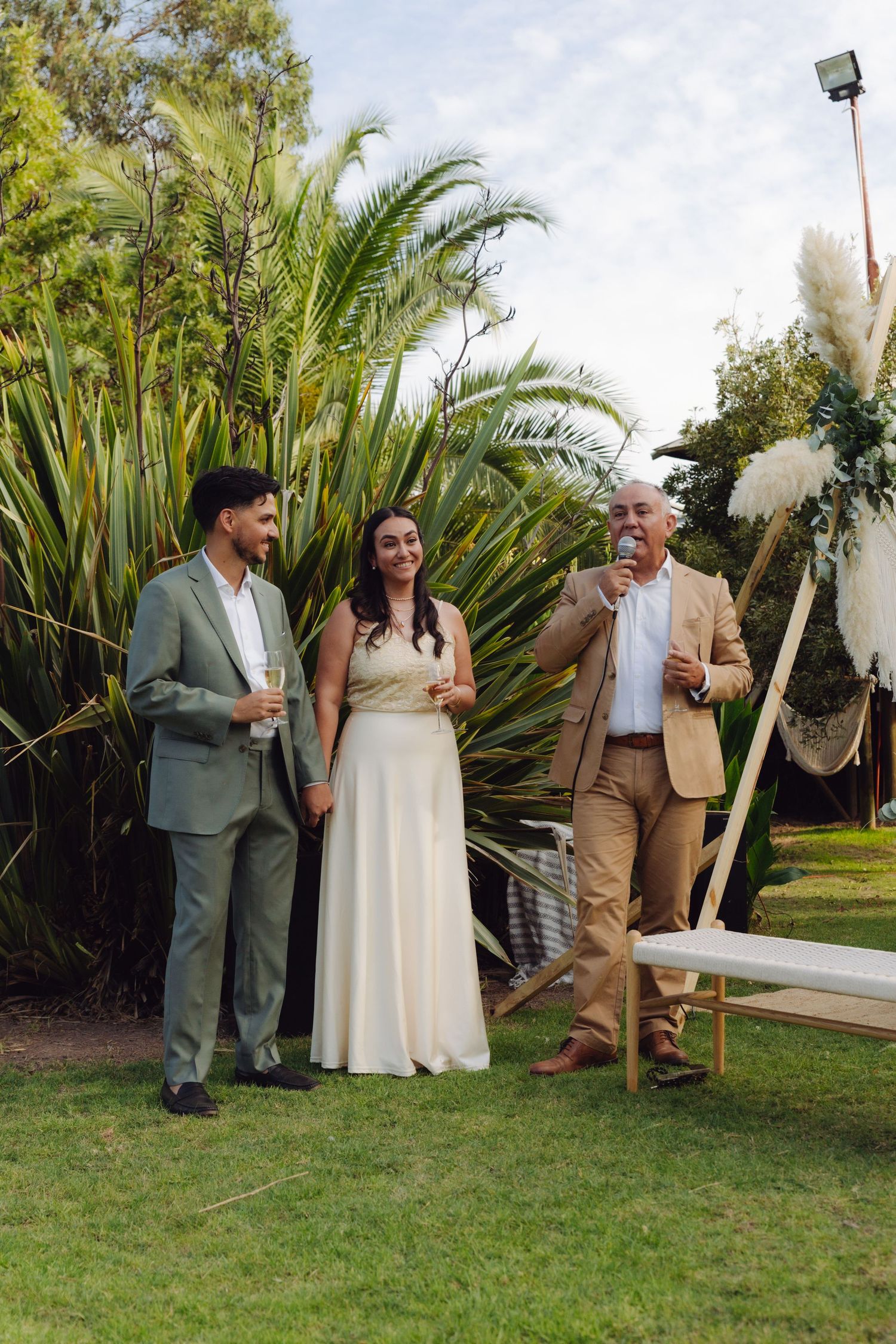 Groom in grey suit and bride in white dress laughing with older man speaking at outdoor wedding toast.