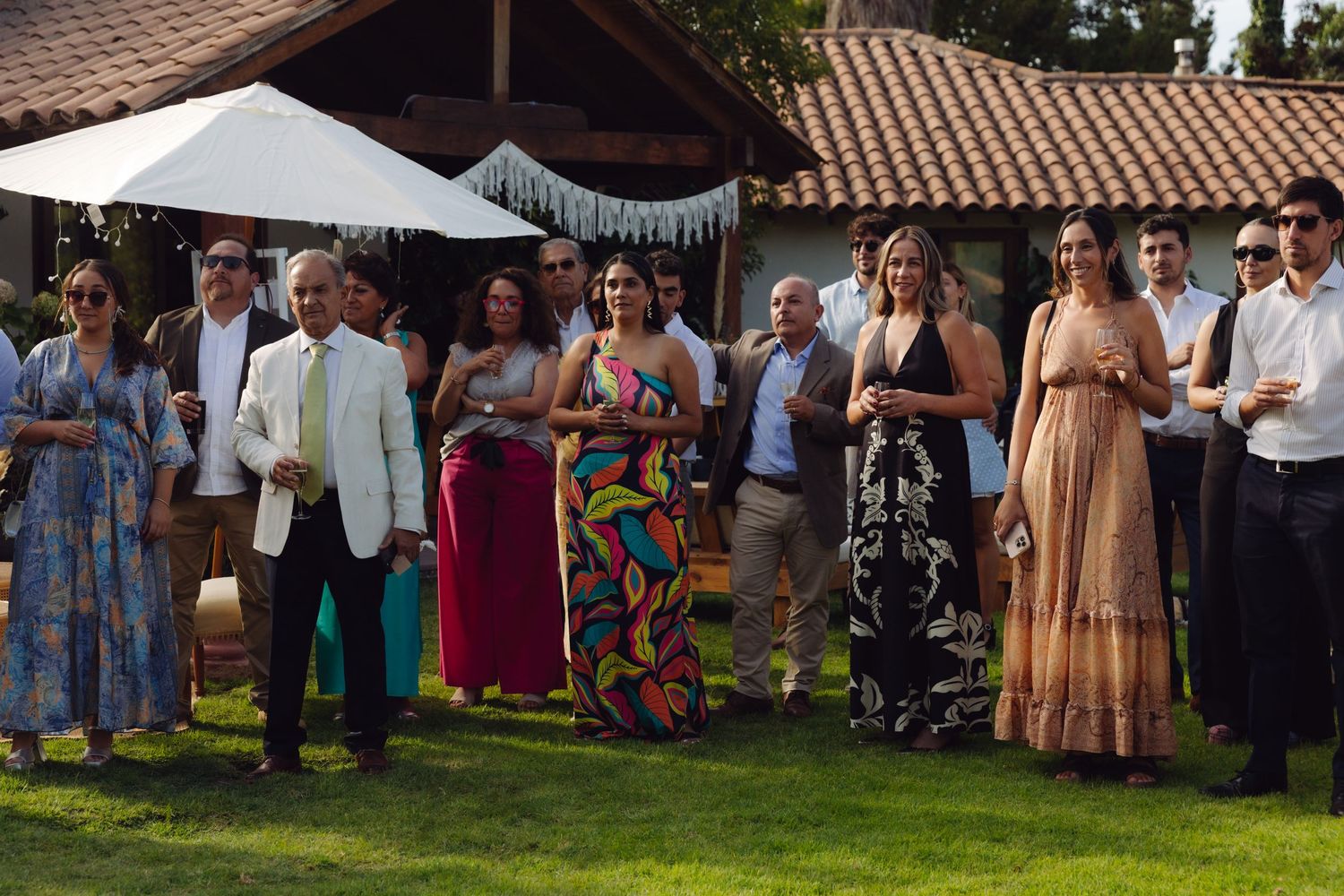 Elegantly dressed wedding guests in colorful gowns gathered on green lawn at outdoor garden wedding.