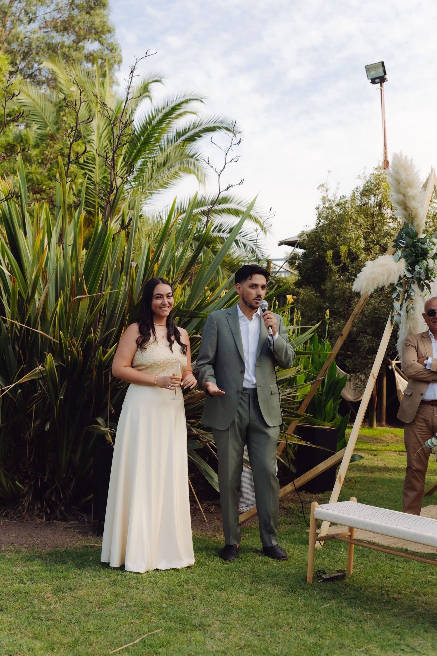 Bride and groom stand together outdoors during wedding ceremony surrounded by tropical greenery.