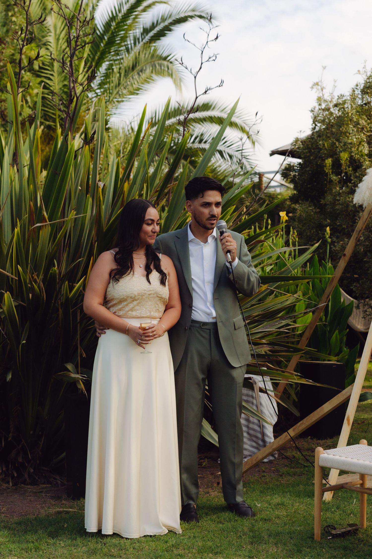 Groom in grey suit speaking into microphone beside bride in white dress at lush outdoor garden wedding.