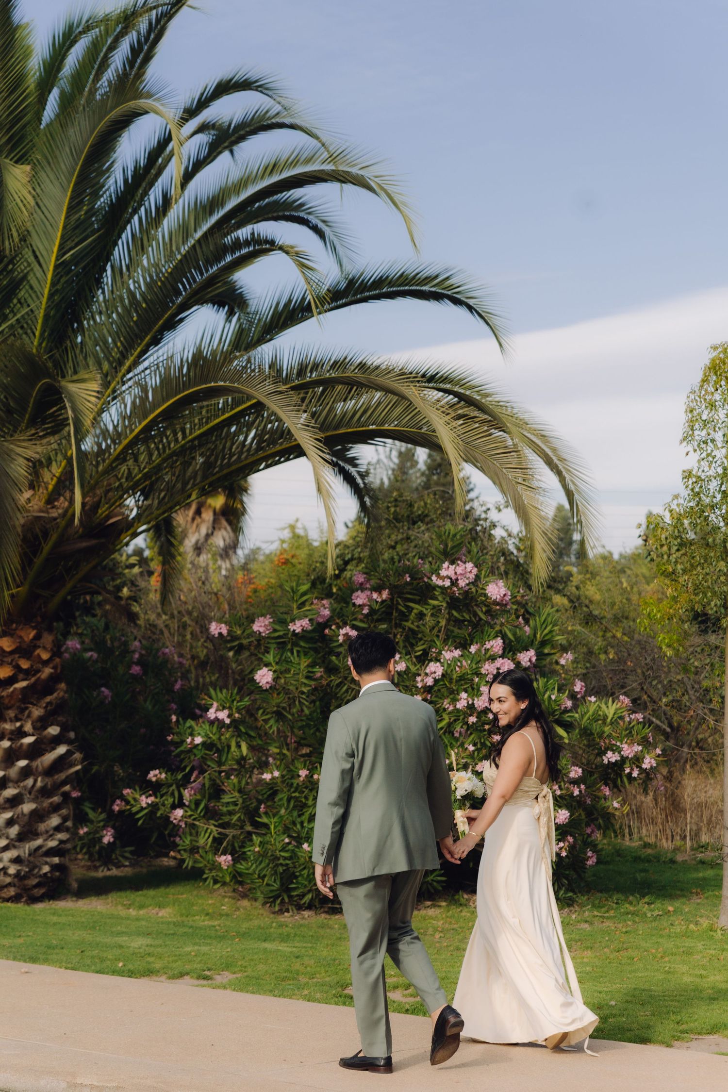 Newlywed couple walks hand in hand along garden path with blooming pink flowers and palm tree.