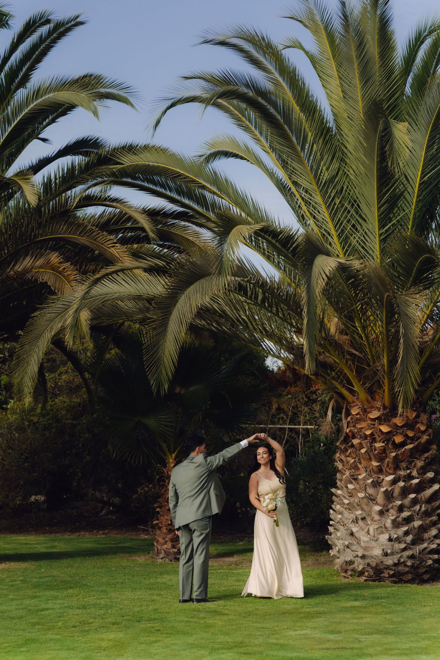 Groom twirls bride in elegant white gown on green lawn beneath large tropical palm trees.