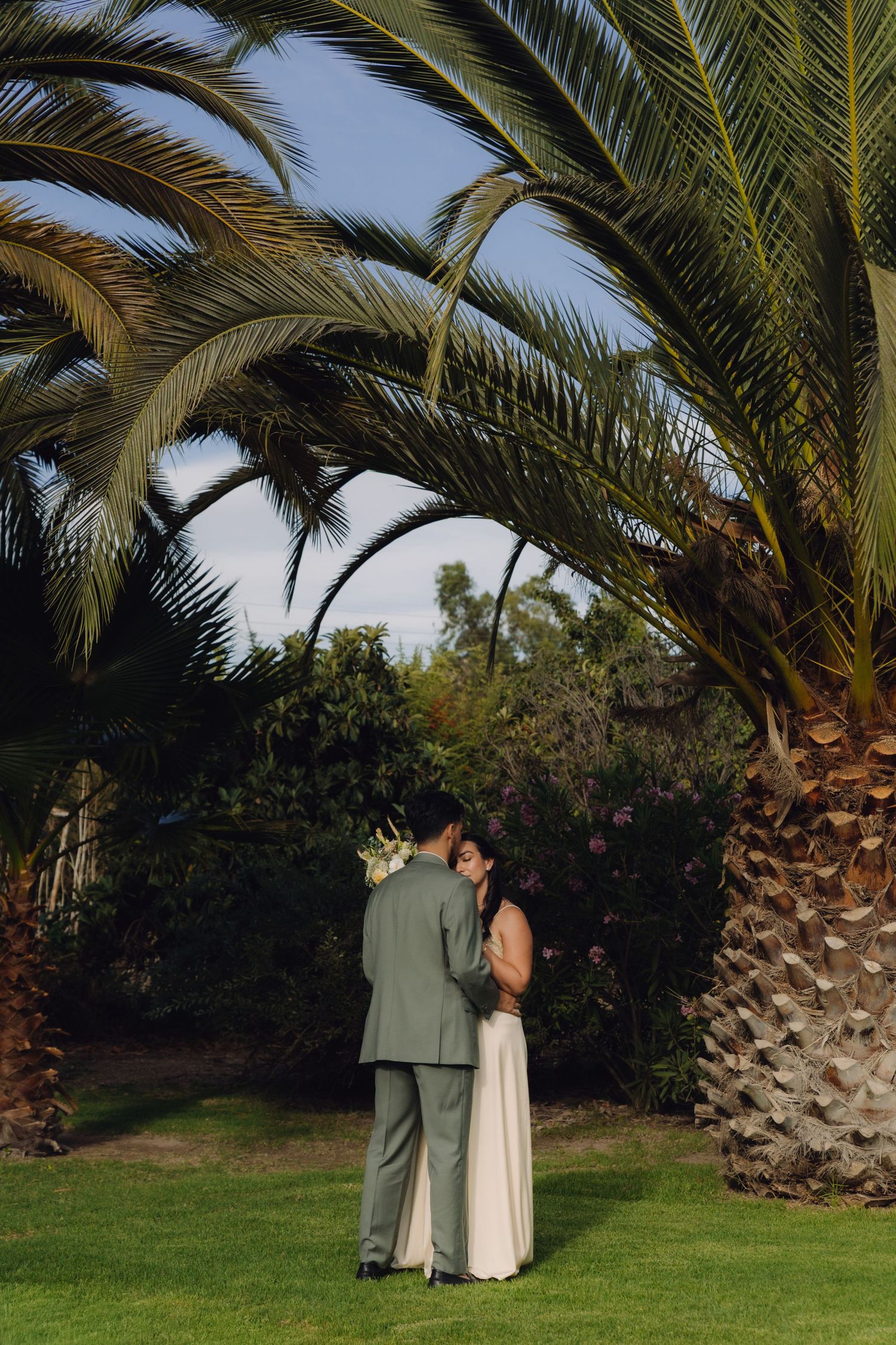 Bride and groom share intimate embrace under sweeping palm tree fronds in lush garden setting.