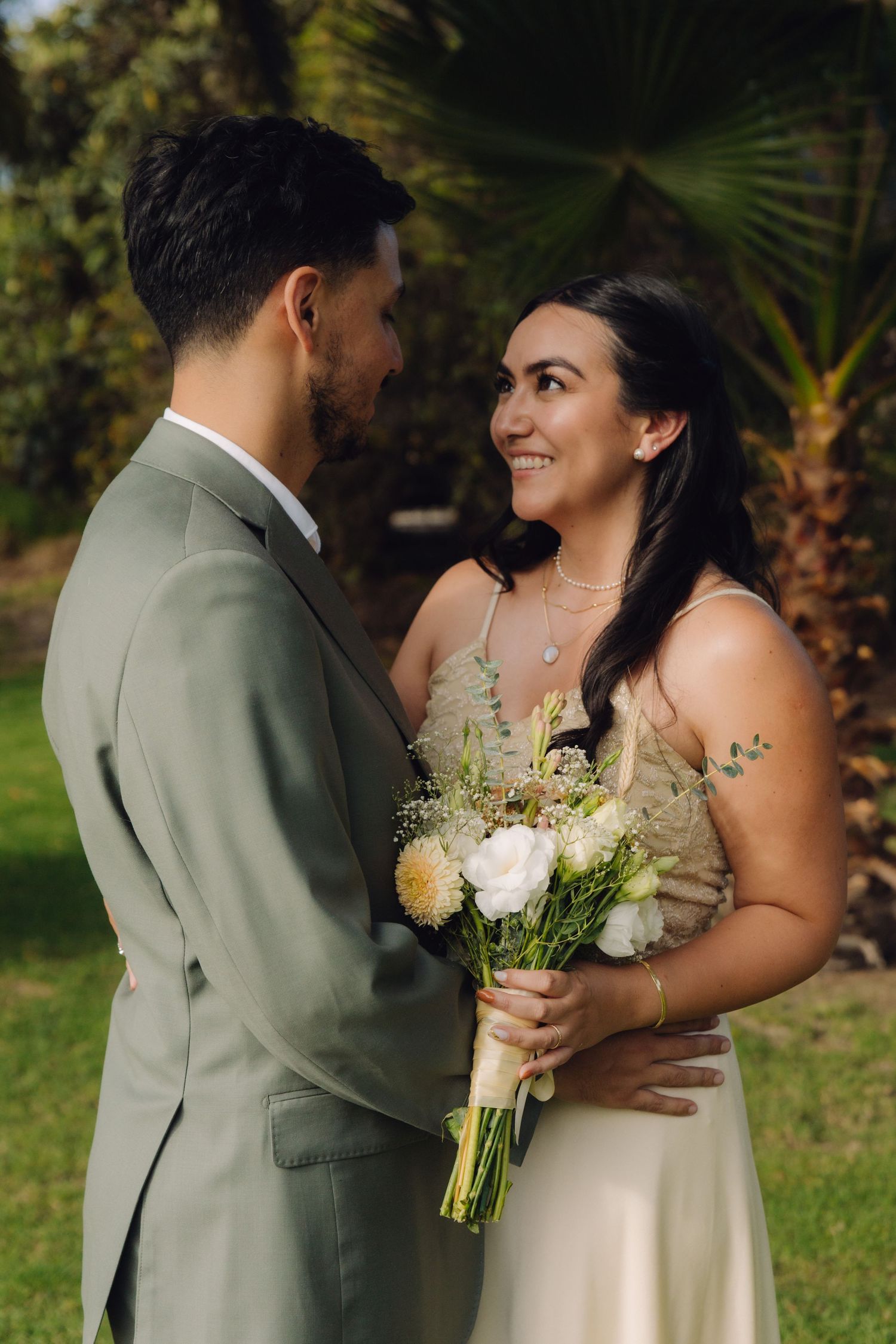 Smiling bride holds white and cream bouquet while gazing at groom in sage green suit outdoors.