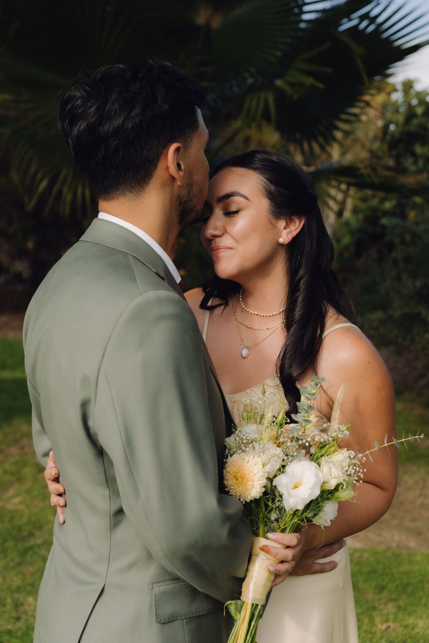 Bride with eyes closed leans close to groom while holding white floral bouquet in tropical garden.