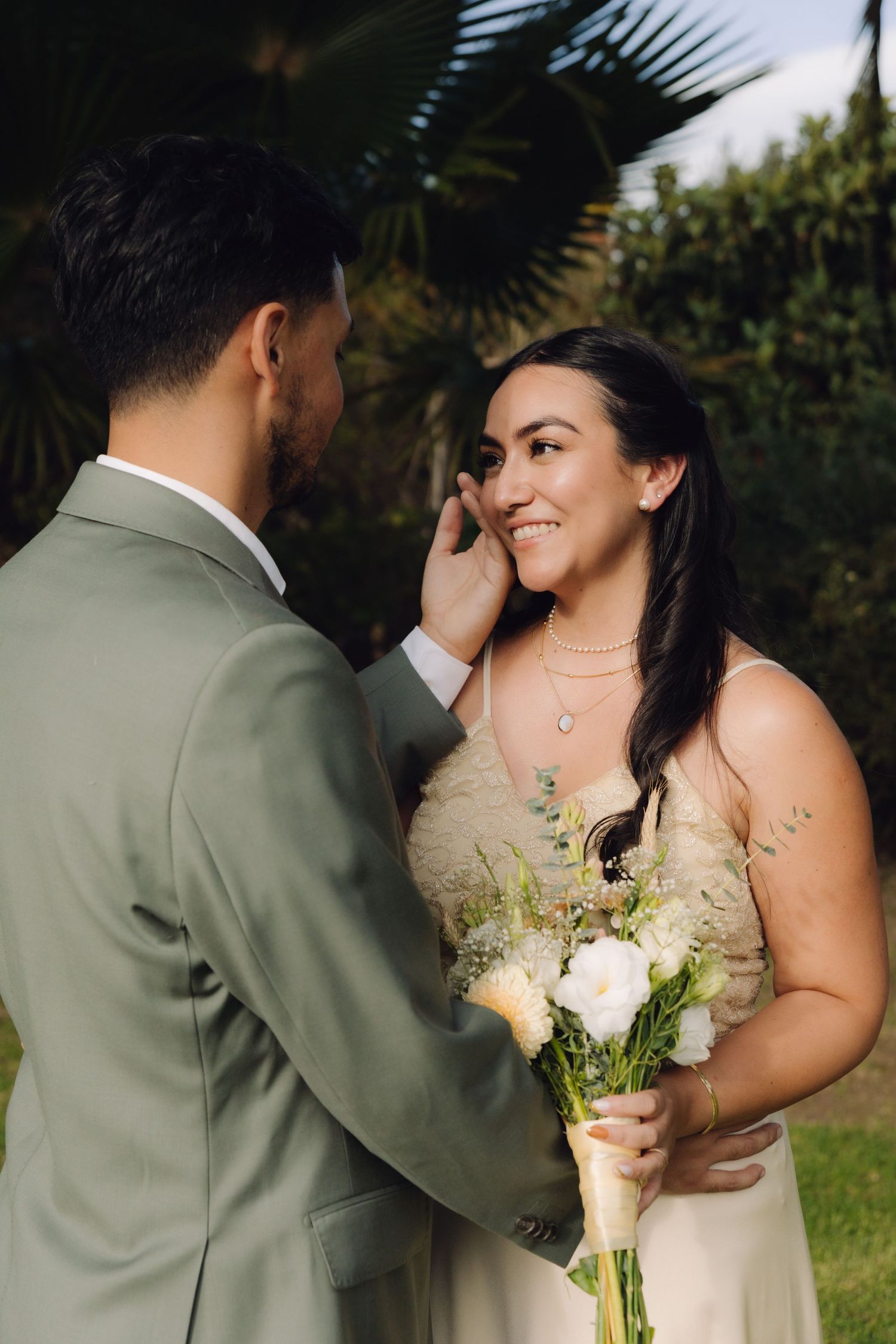 Groom gently touches brides face as she smiles holding bouquet in sun-dappled garden setting.