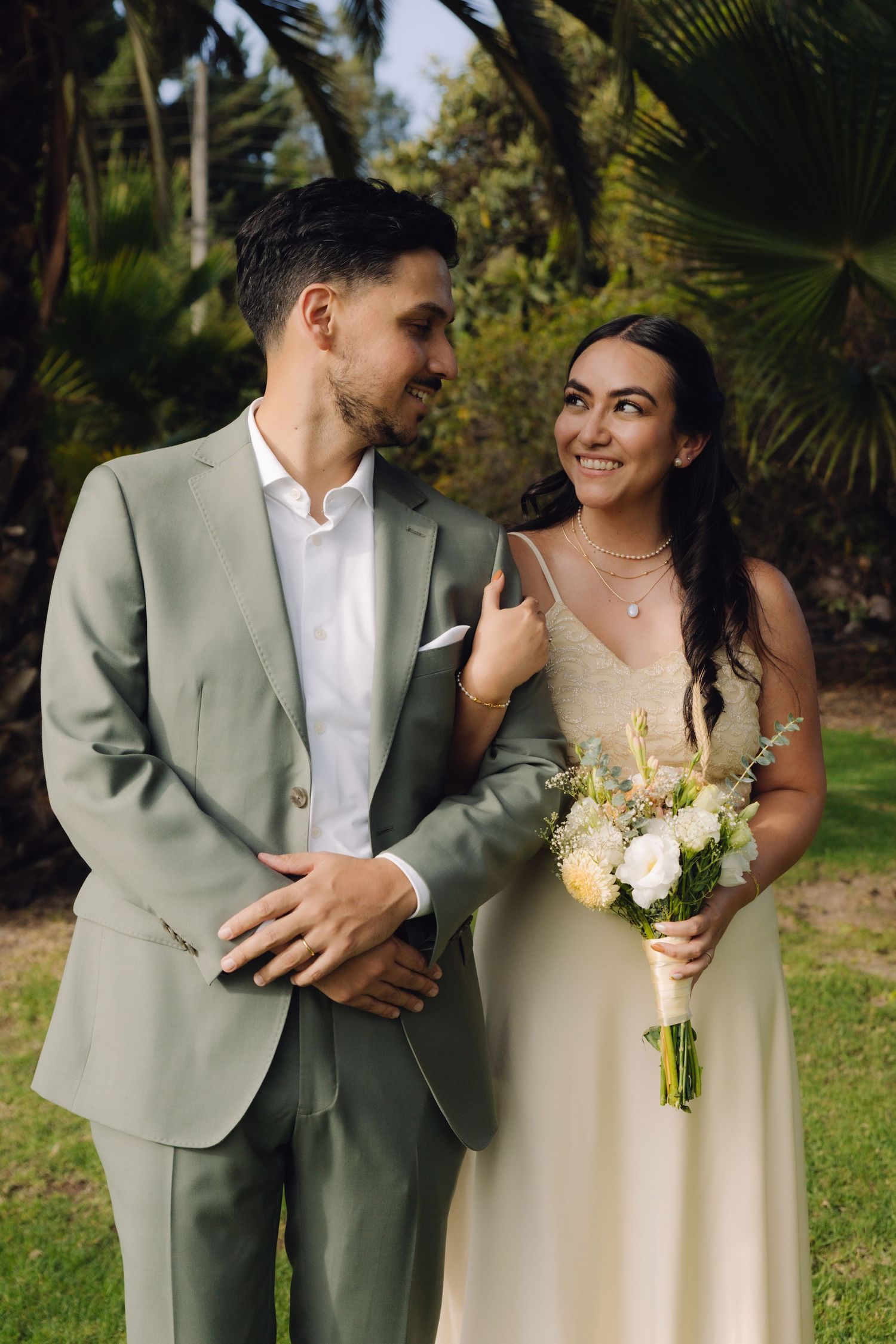 Bride in cream gown holds bouquet and smiles at groom in sage suit amid lush green garden.