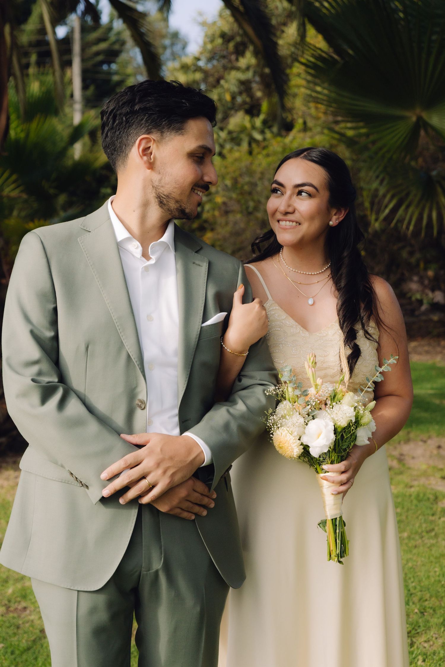 Elegant couple poses together in tropical garden as bride holds white and peach wedding bouquet.