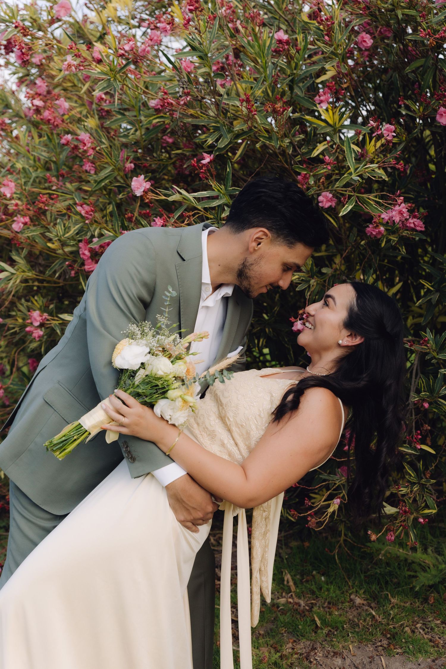 Wedding couple sharing an intimate moment in front of vibrant pink oleander flowers, bride holding bouquet.
