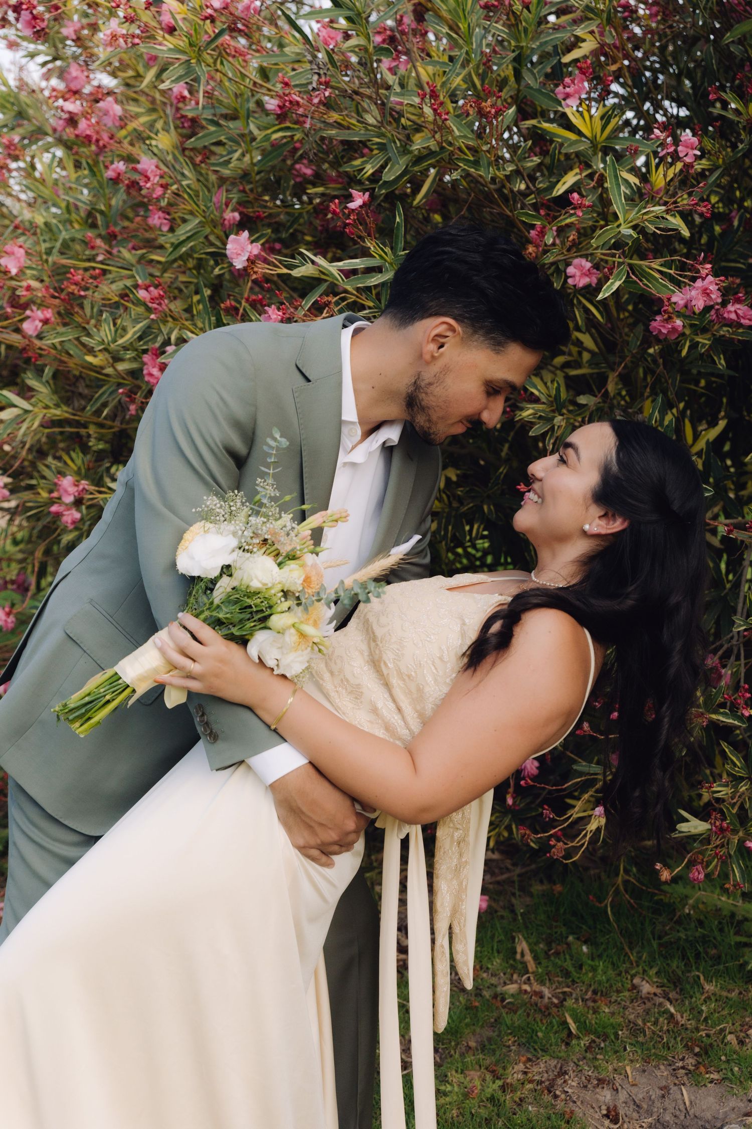 Bride and groom sharing a romantic dip kiss in front of pink flowering bushes, bride holding white bouquet.