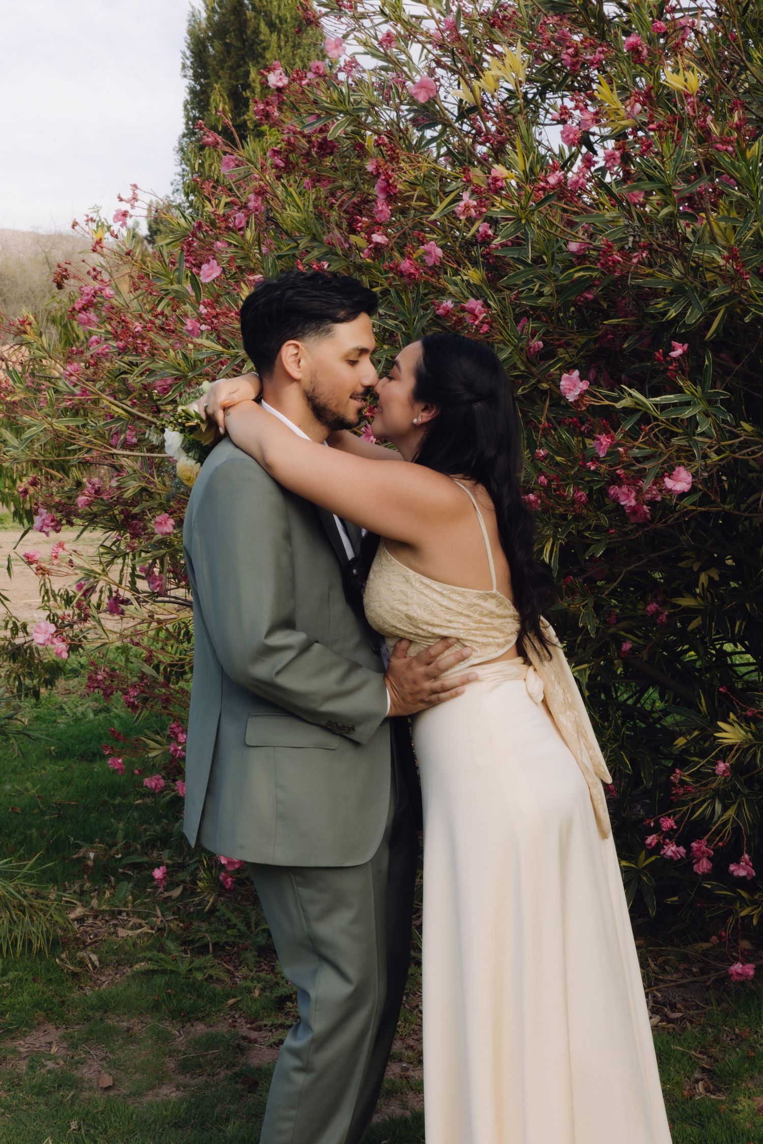 Bride and groom kissing tenderly in a garden setting surrounded by lush pink flowering oleander bushes.