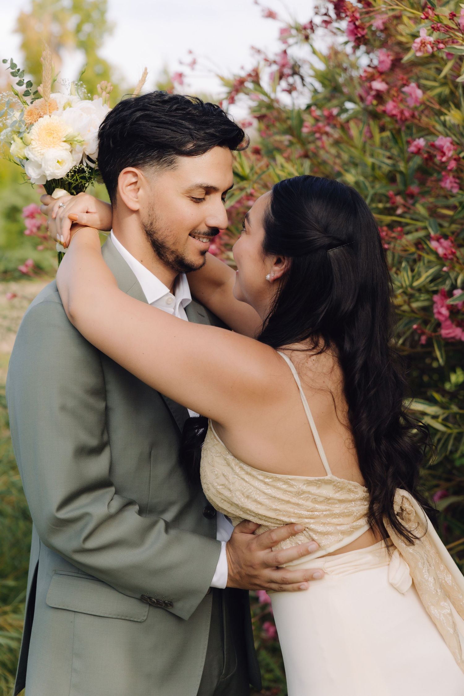 Smiling bride and groom embracing closely in front of pink blooming flowers during outdoor wedding photos.