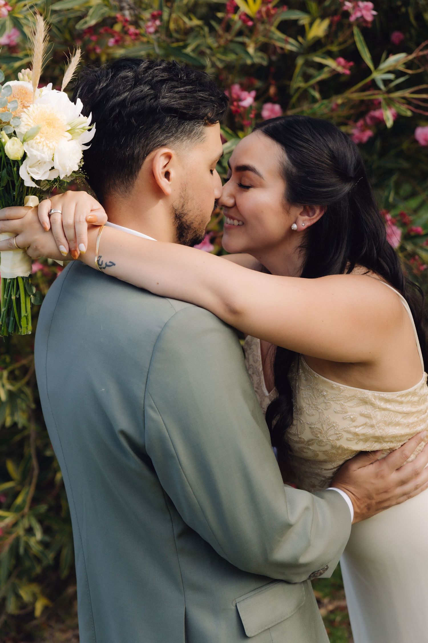 Bride and groom sharing a romantic kiss surrounded by pink flowering bushes on wedding day.