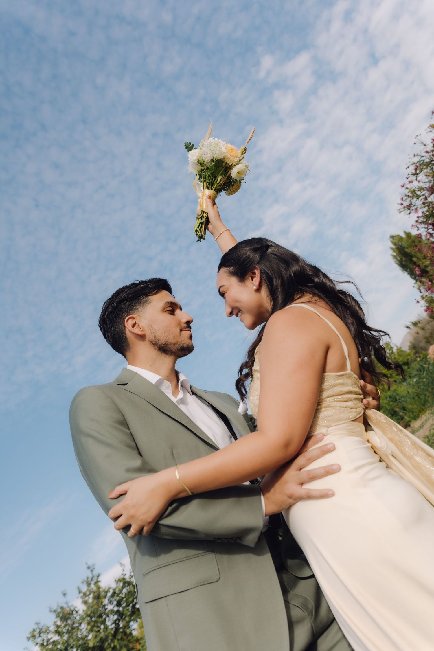 Groom lifting bride as she raises bouquet against blue sky in joyful wedding celebration.