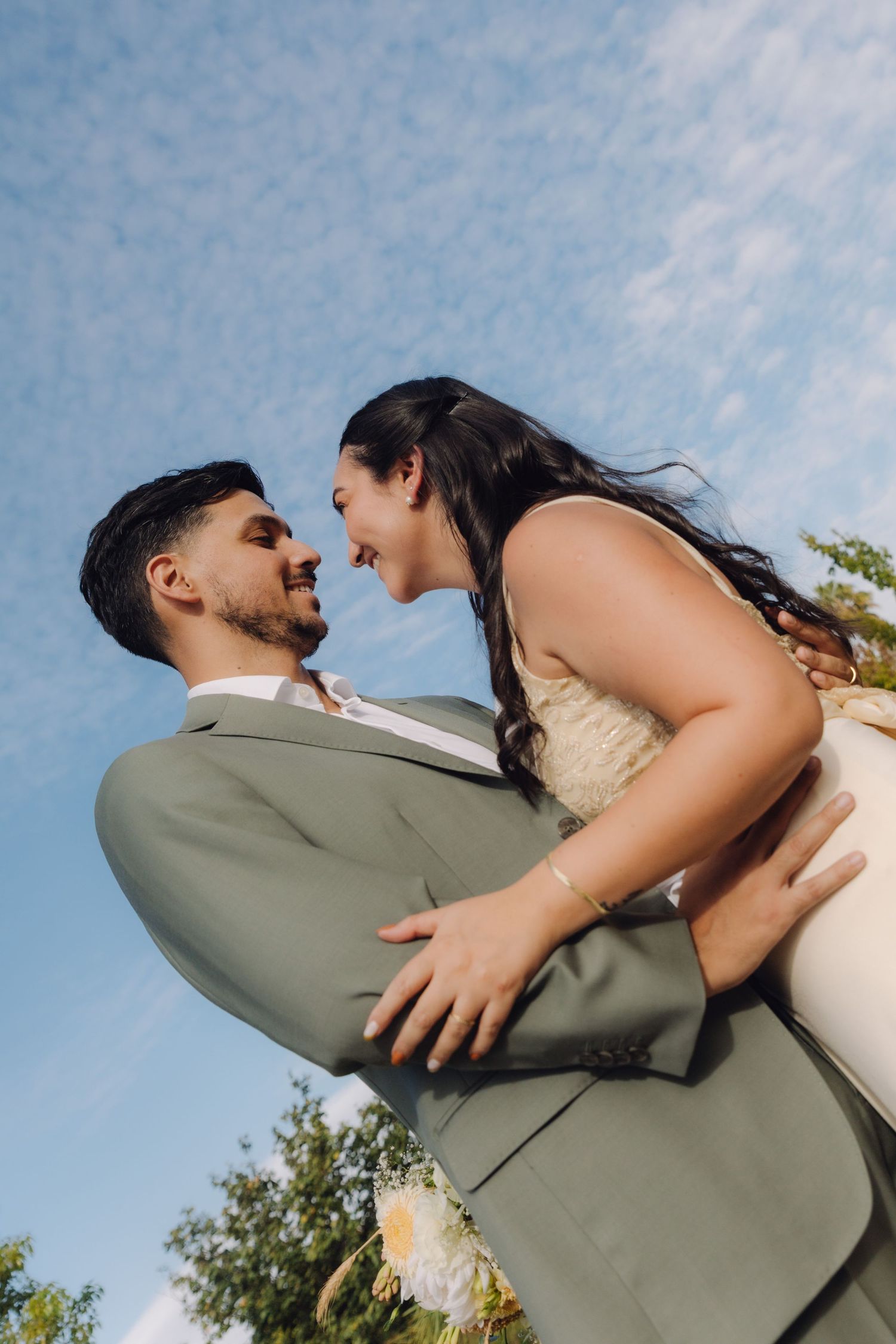 Groom lifting smiling bride against blue sky, both gazing lovingly at each other outdoors.
