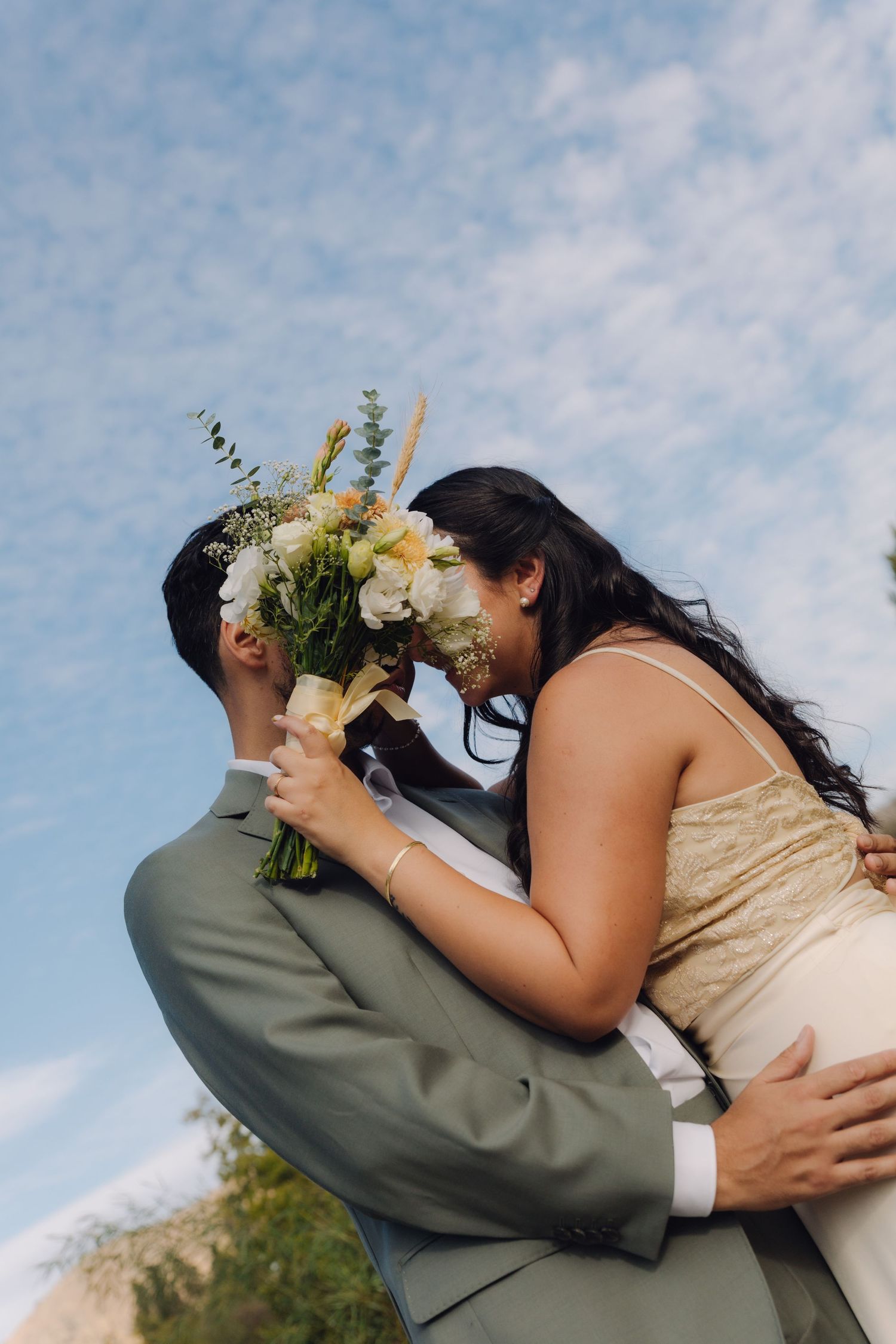 Groom lifting bride as she leans in for a kiss holding white wedding bouquet outdoors.