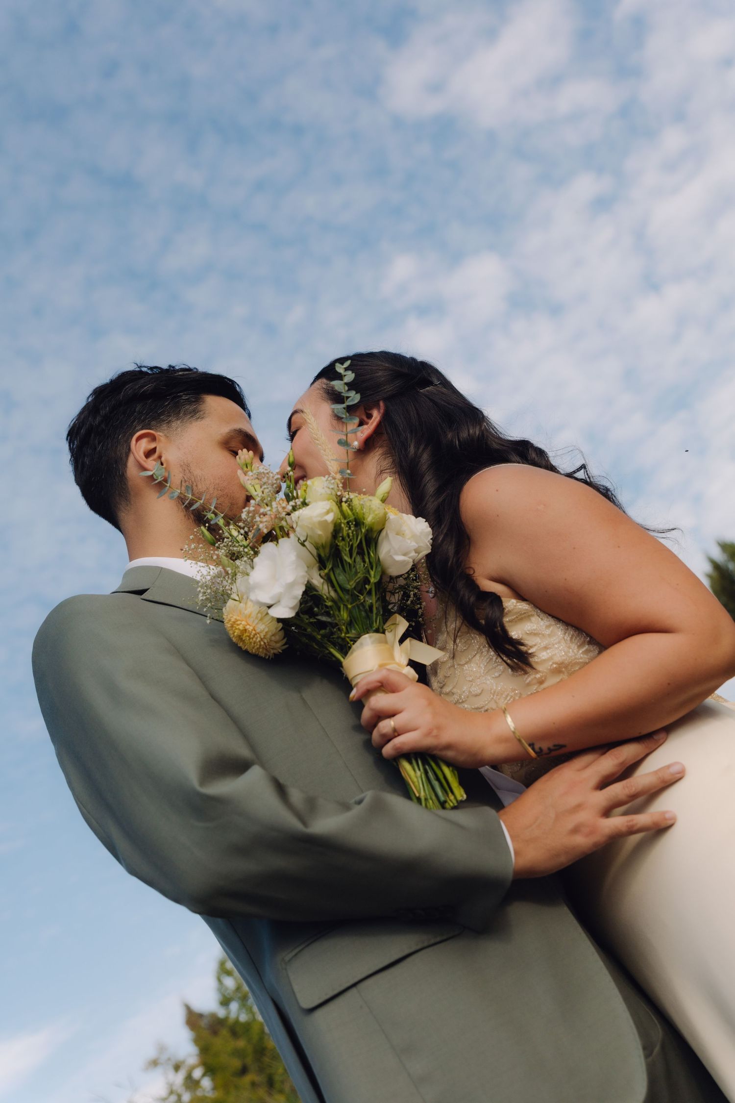 Bride and groom sharing a tender kiss while holding white floral bouquet under blue sky.