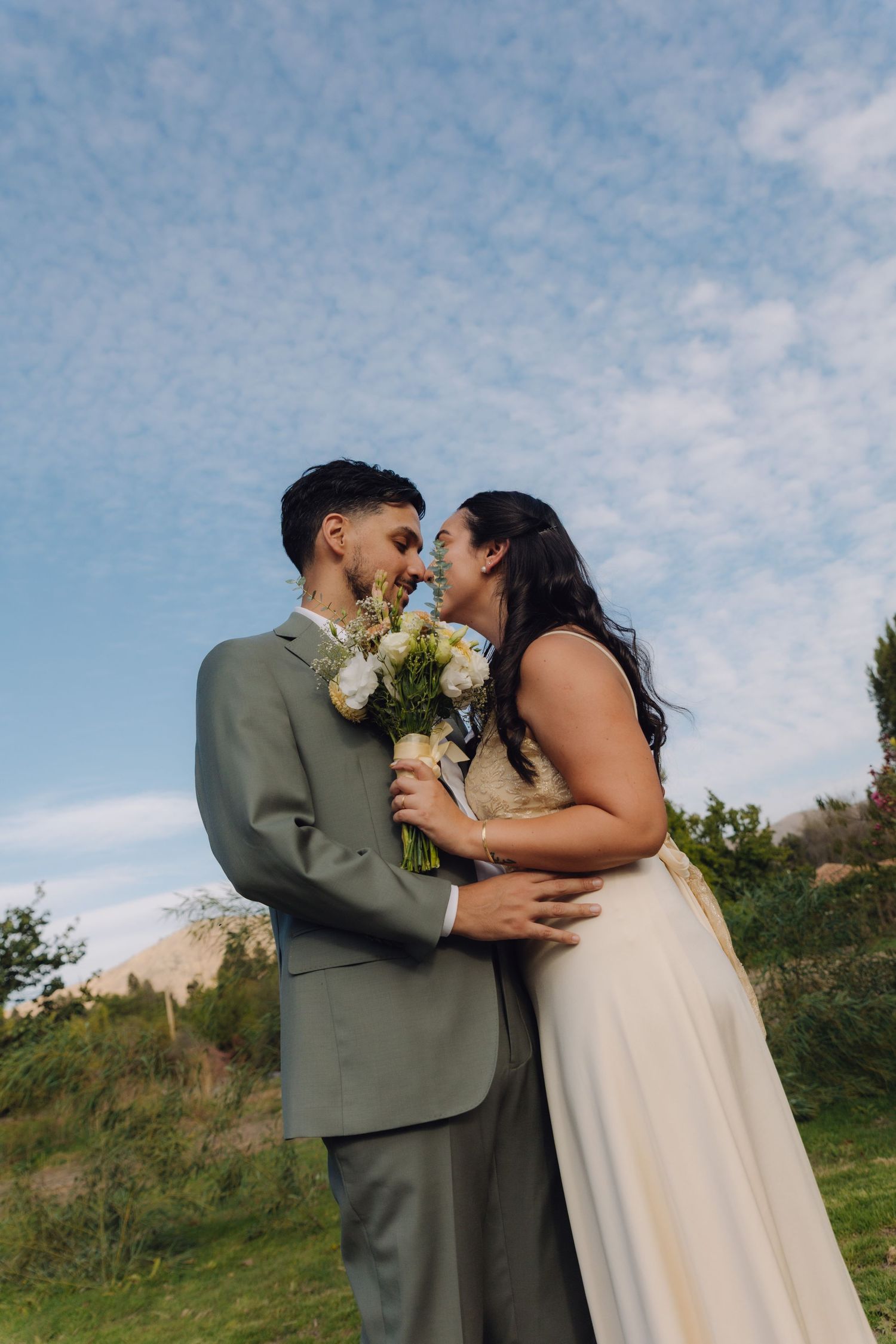 Bride and groom sharing an intimate moment with white bouquet against a scenic blue sky.