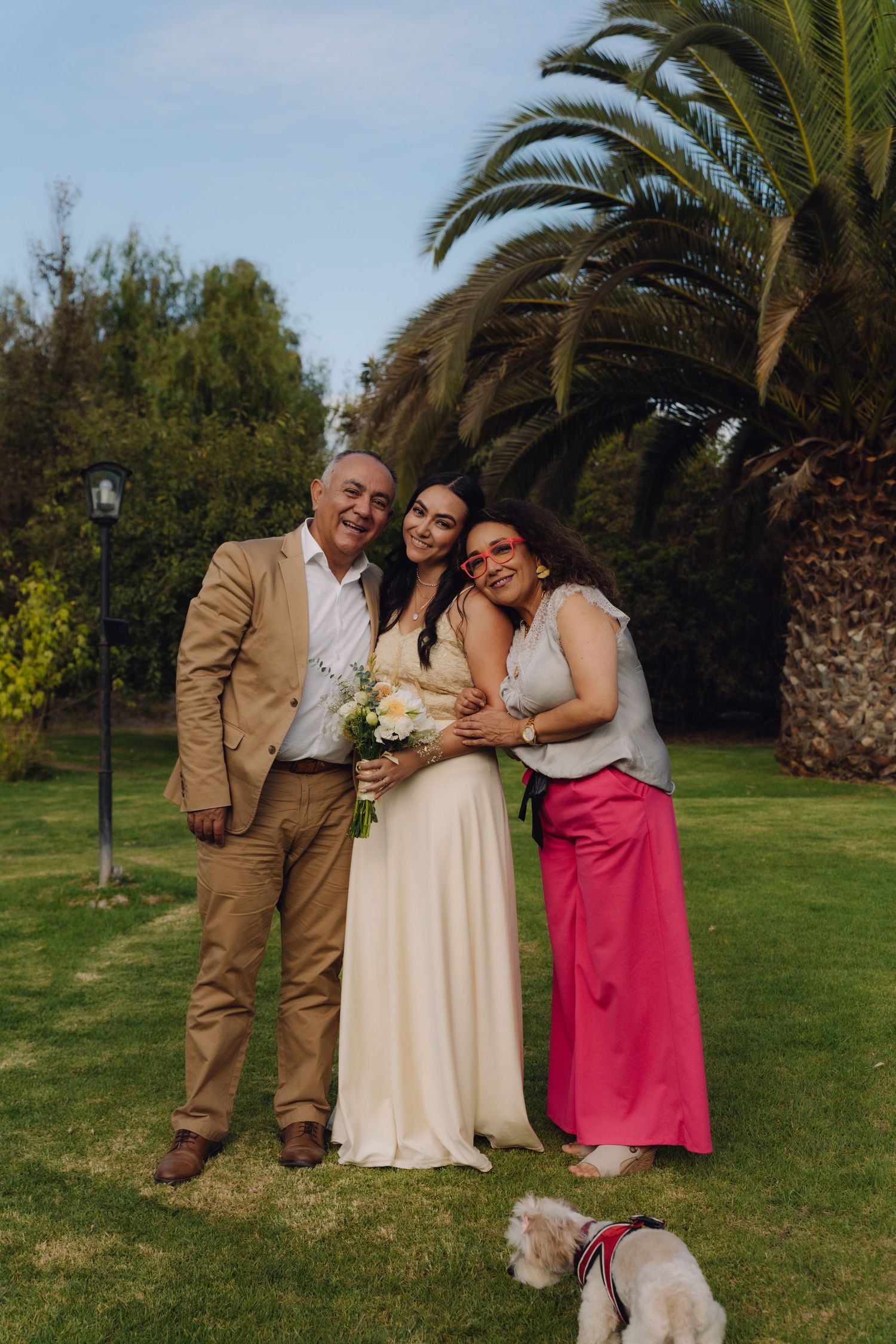 Bride posing with parents and small dog on green lawn with tall palm tree backdrop.