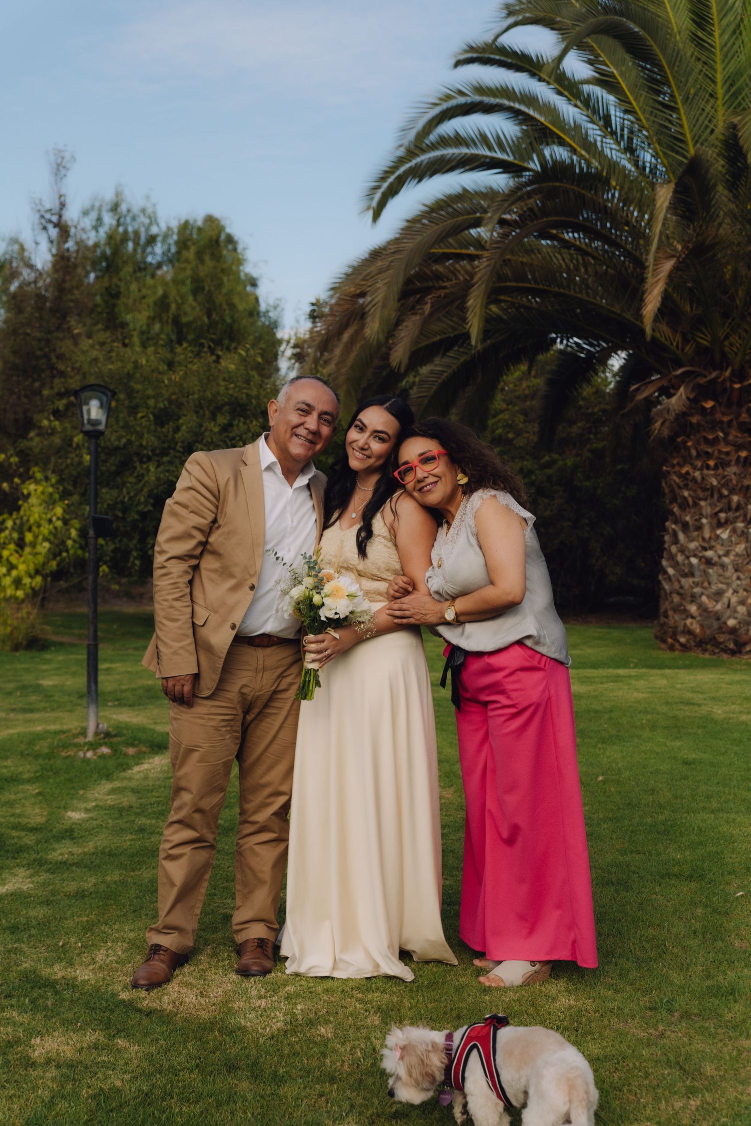 Bride smiling with parents on lush green lawn with large palm tree in background.