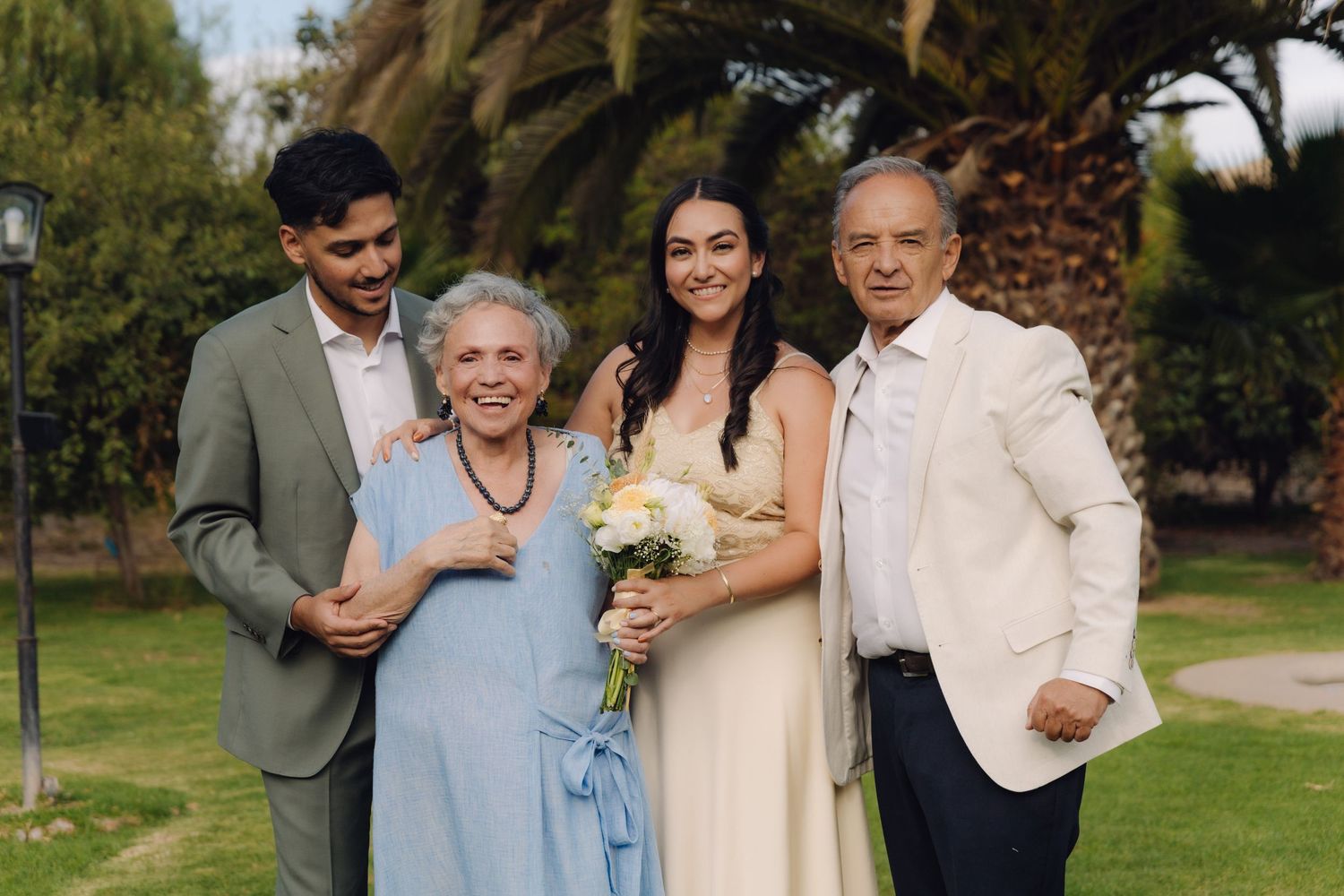 Bride with bouquet poses with family including elderly couple at outdoor wedding ceremony.