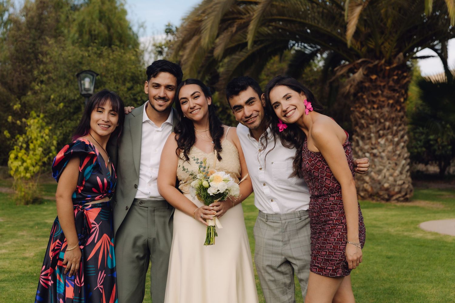 Bride holding bouquet smiles with four friends at outdoor wedding celebration.
