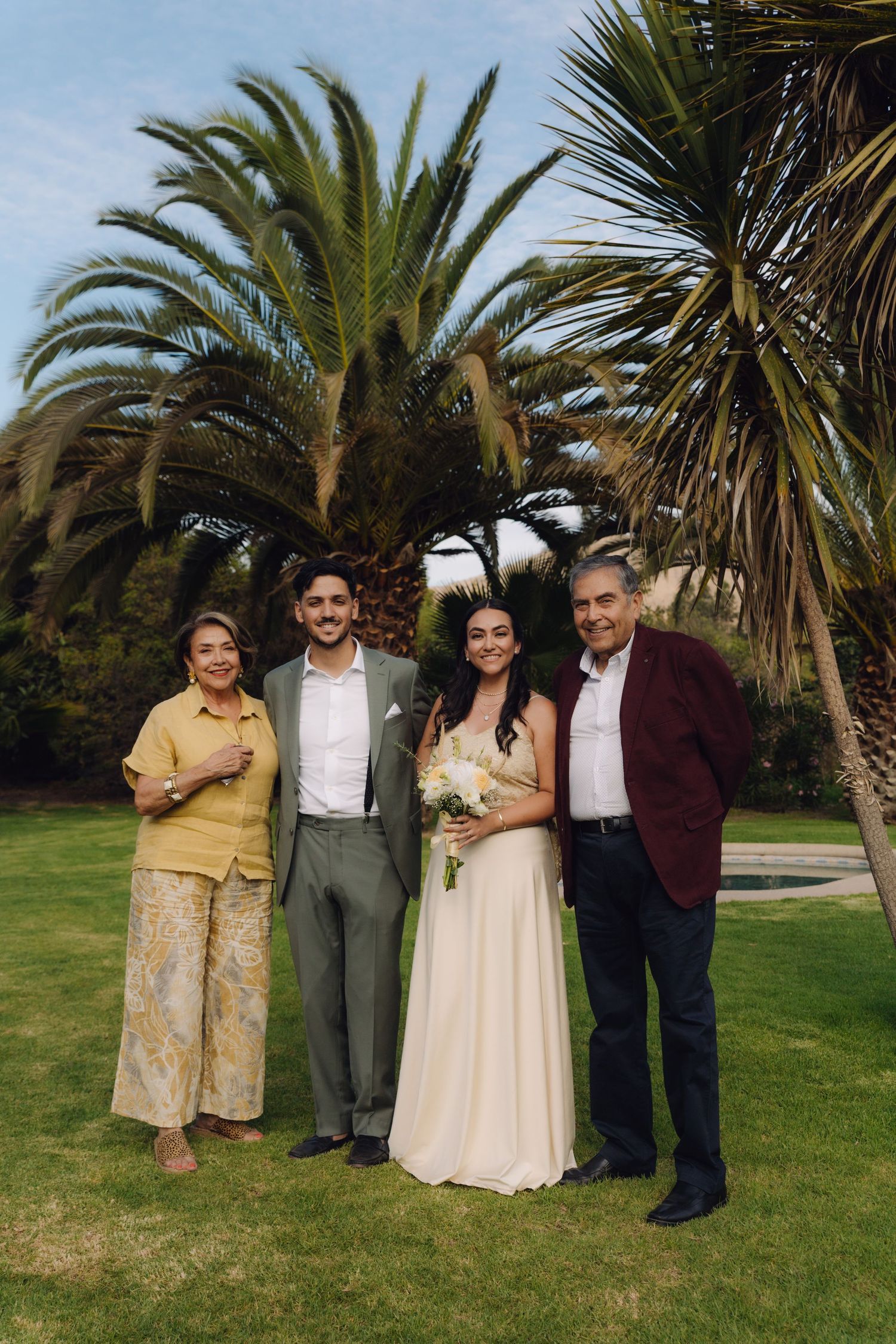 Bride and groom pose with parents under palm trees at outdoor wedding.