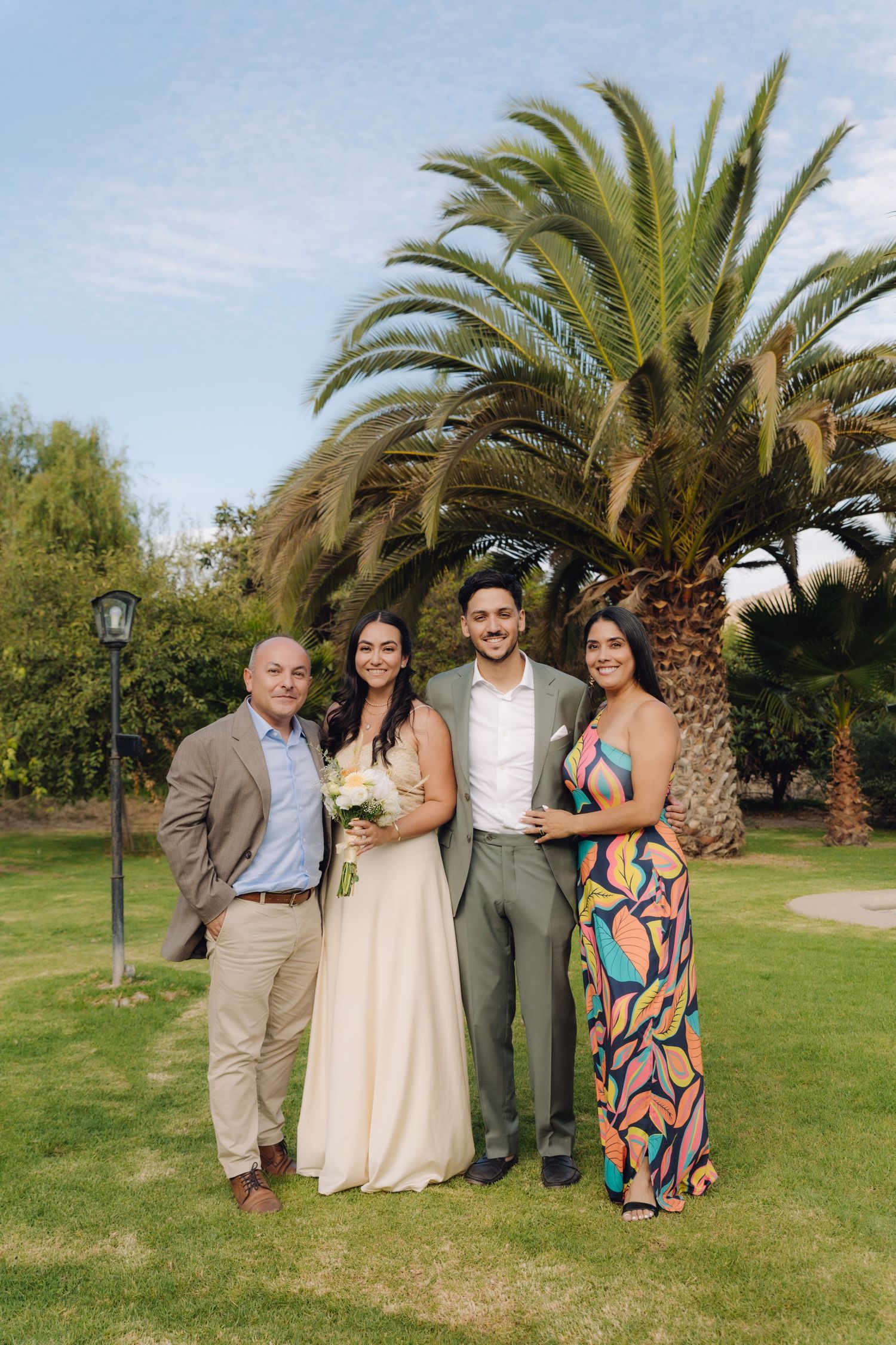 Wedding couple poses with two guests in colorful attire on green lawn.