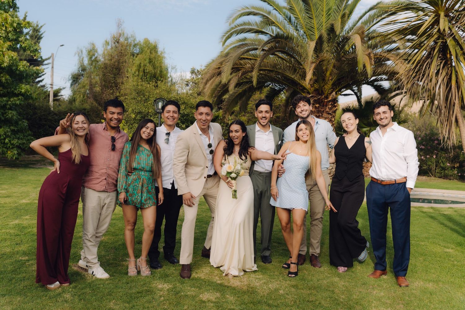 Large group of wedding guests smile together outdoors near tropical palm trees.