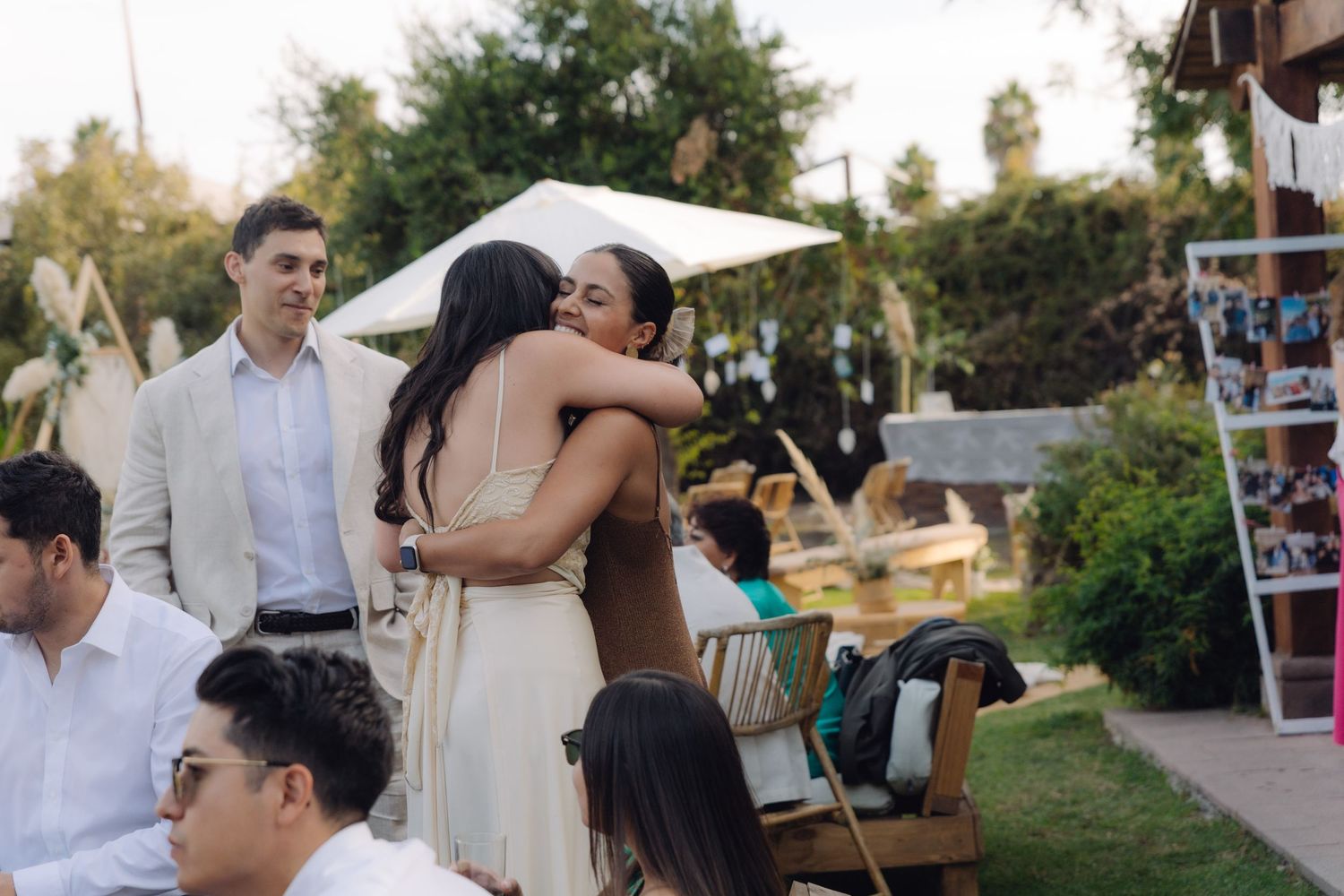 Two women share a warm embrace at an outdoor wedding reception surrounded by guests.