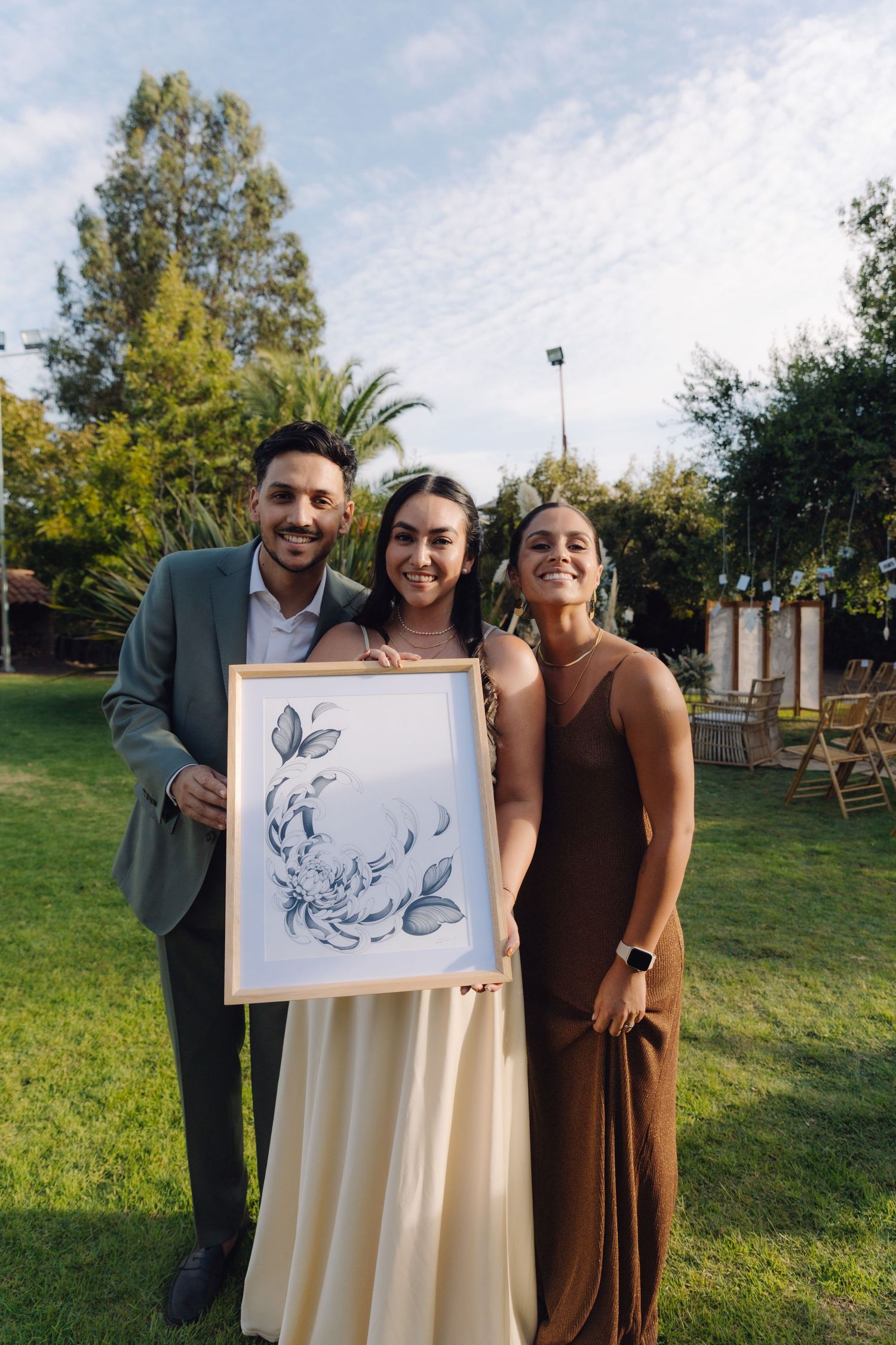 Smiling couple and friend hold a floral artwork print at an outdoor wedding celebration.