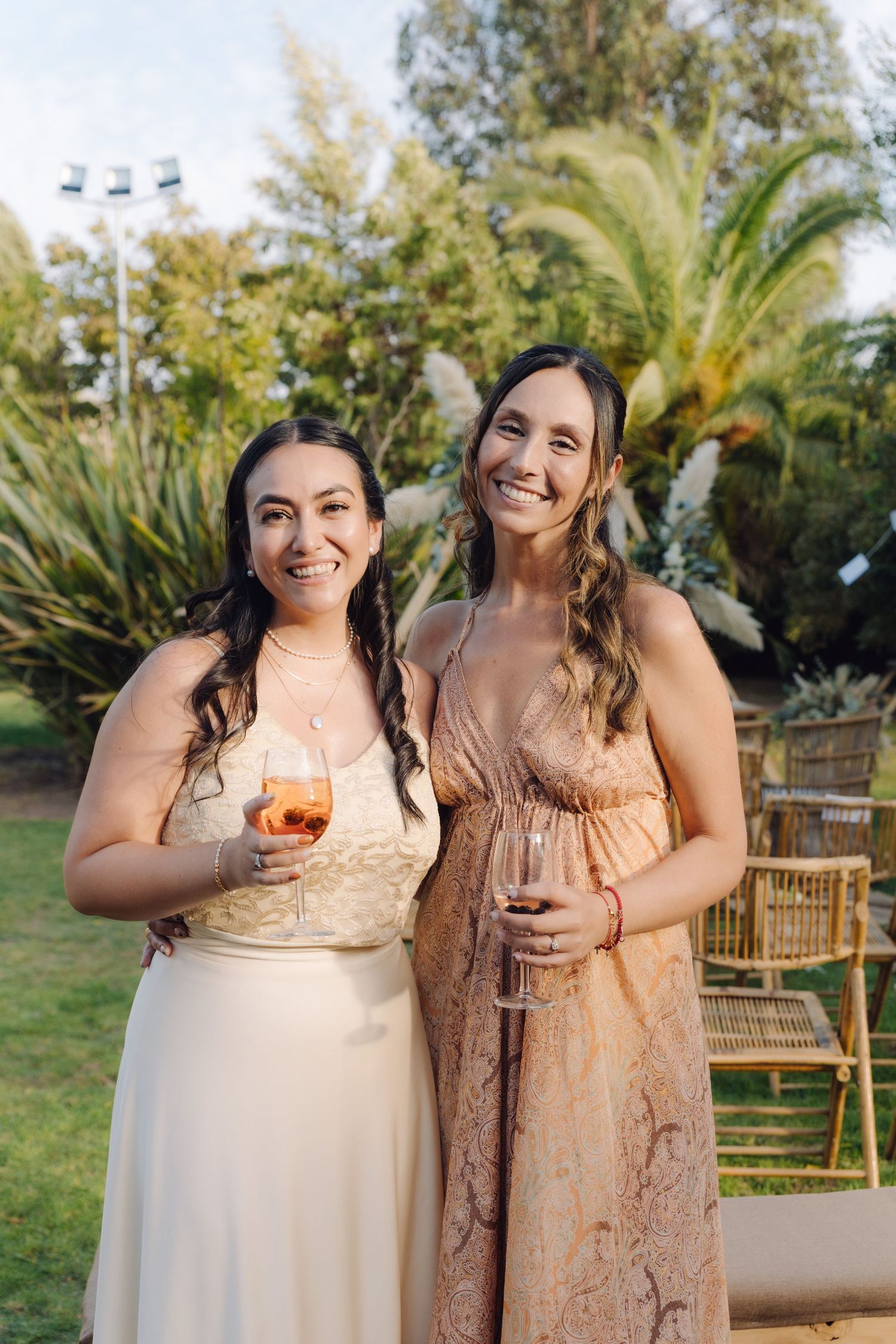 Two women in elegant dresses smile while holding wine glasses at an outdoor wedding.