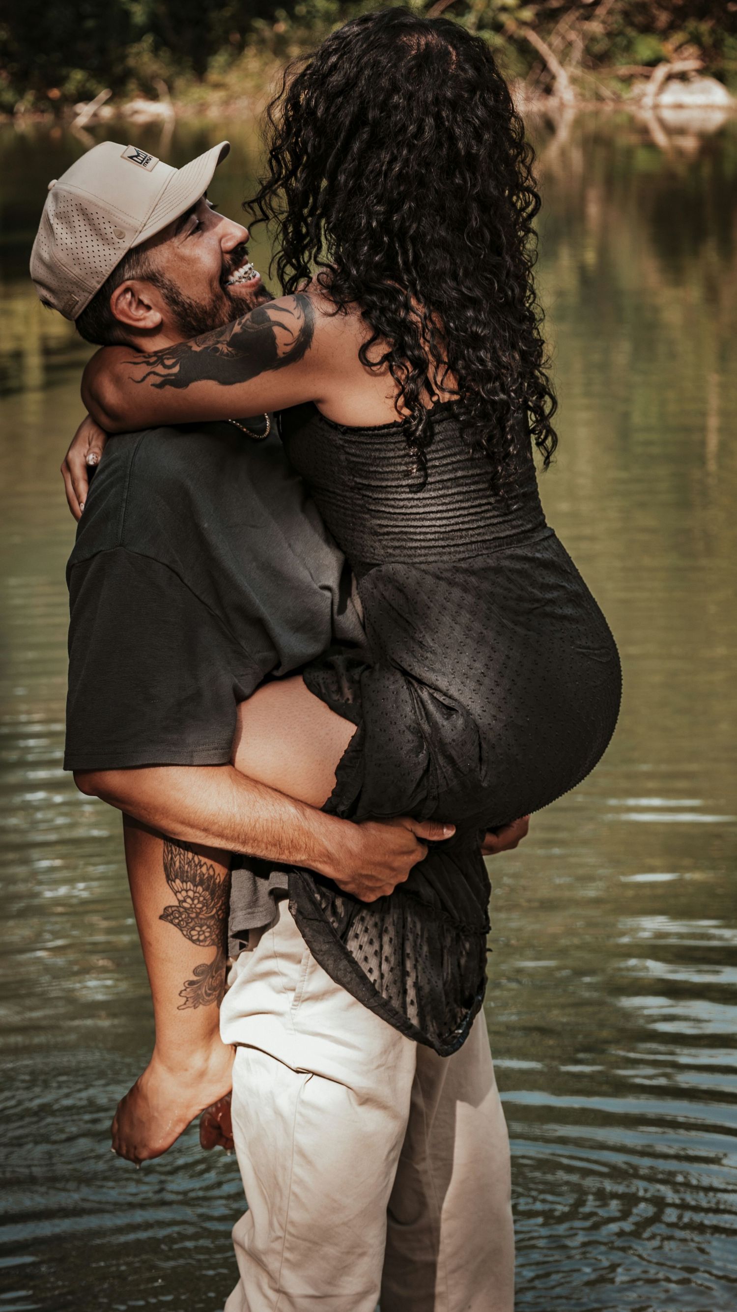 A tattooed man holds a woman in his arms while standing in a river, sharing a romantic moment outdoors.
