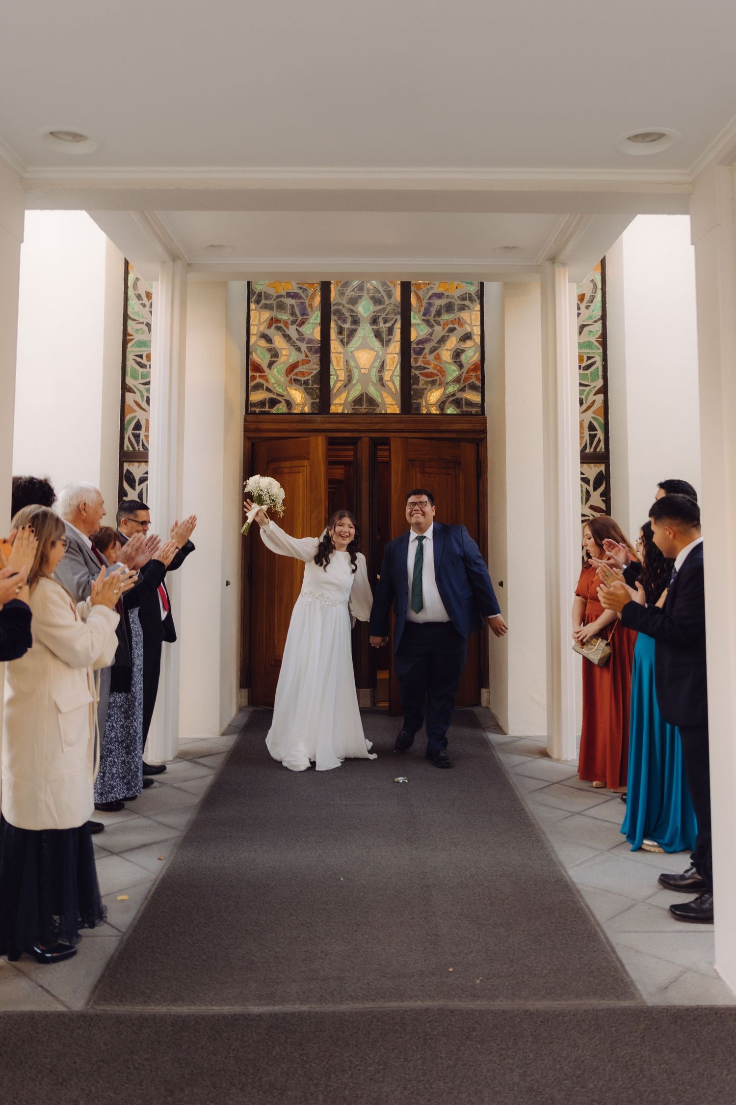 Joyful bride raises bouquet while exiting ceremony venue with groom as guests clap in celebration.