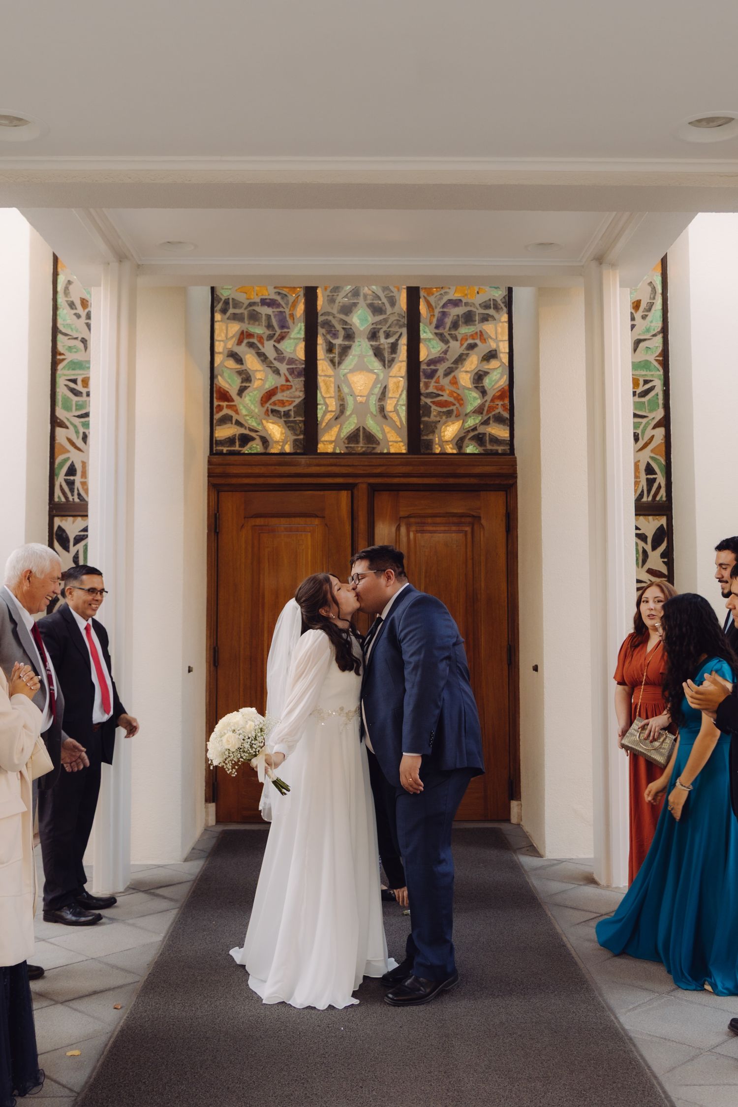 Bride and groom share first kiss at church entrance with colorful stained glass doors and guests watching.