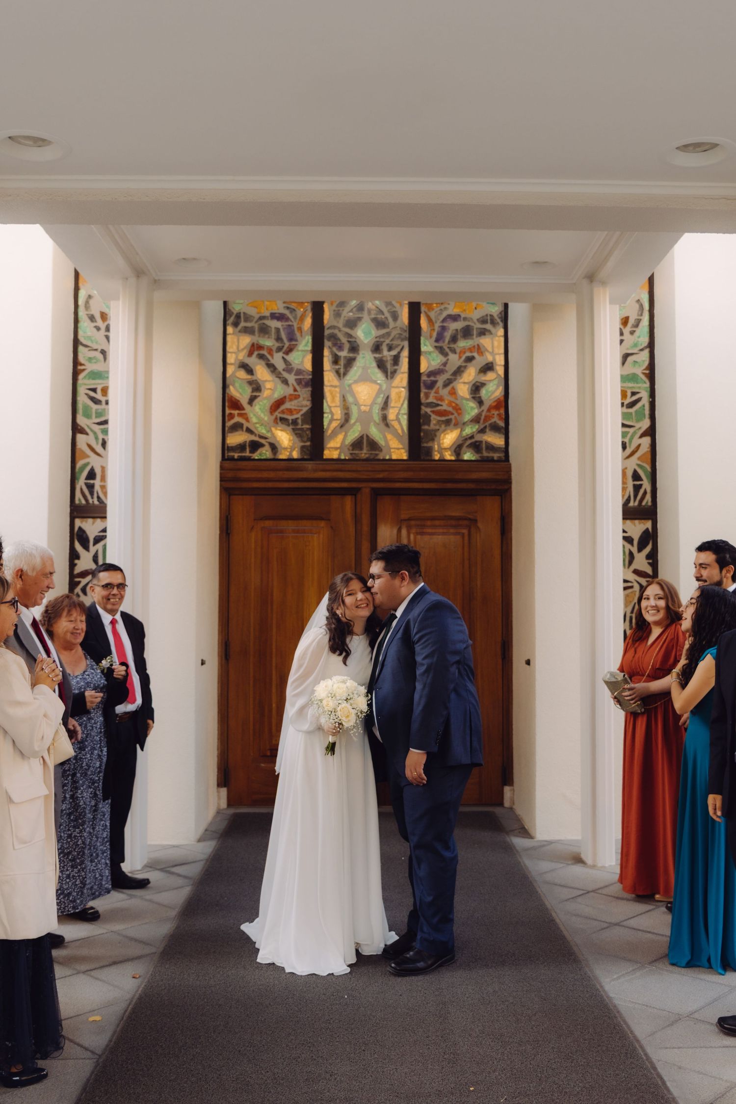 Newlyweds kiss at church entrance flanked by ornate stained glass panels with guests applauding.