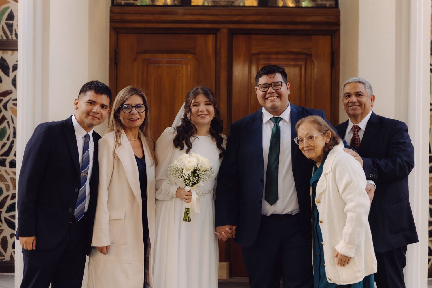 Wedding couple poses with family of six in front of ornate wooden doors at a formal church ceremony.