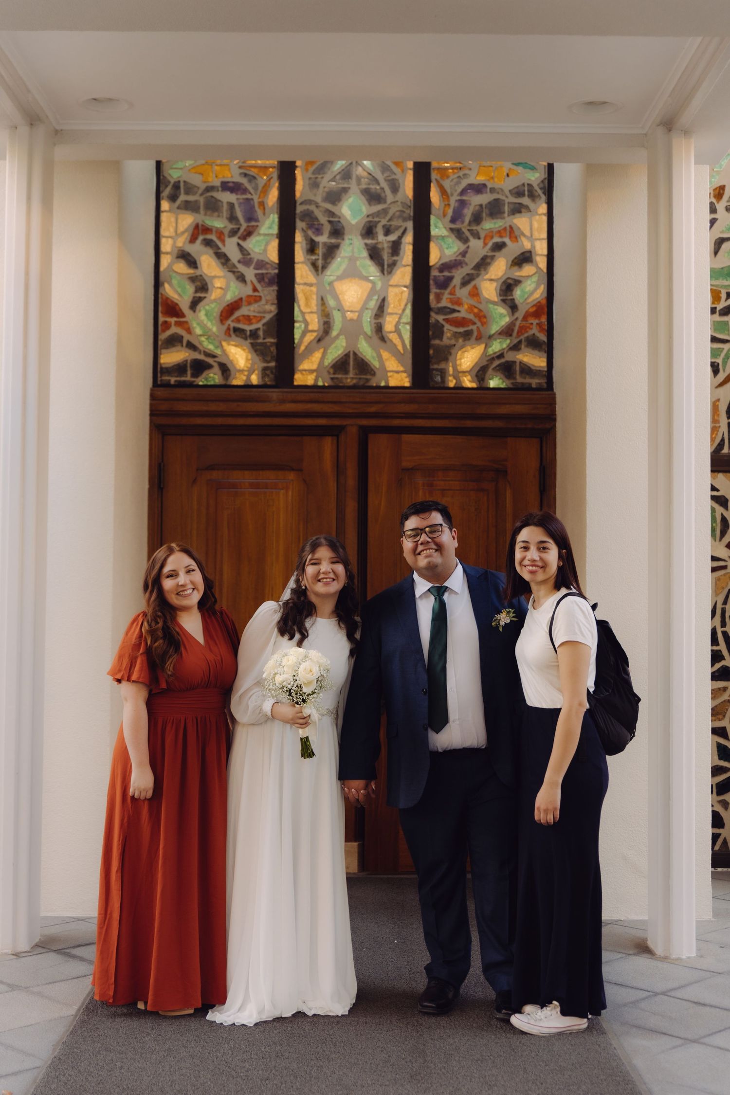 Bride in white gown holds bouquet while posing with three guests before stained glass church doors.