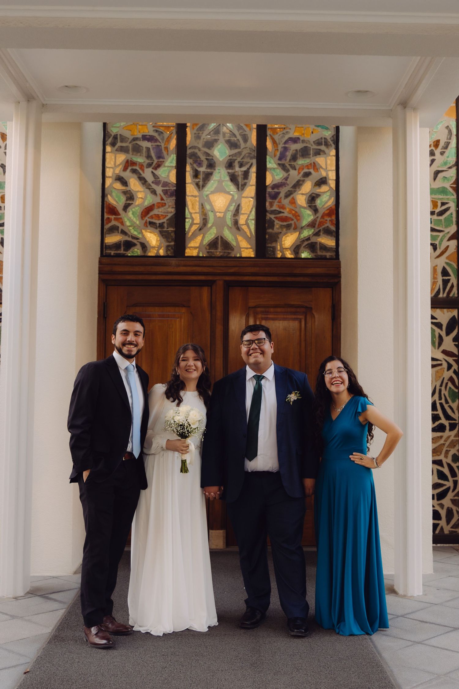 Wedding couple and two guests smile before colorful stained glass windows at a church entrance.