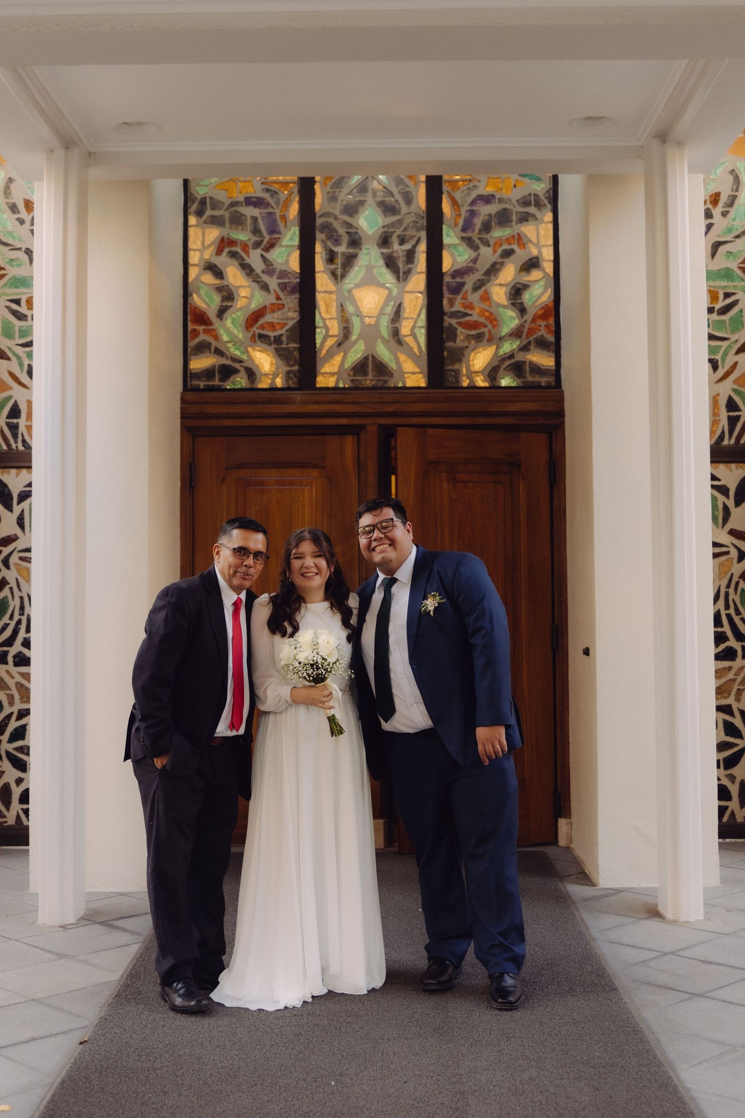 Bride with white bouquet stands between two suited men at a church entrance with stained glass.