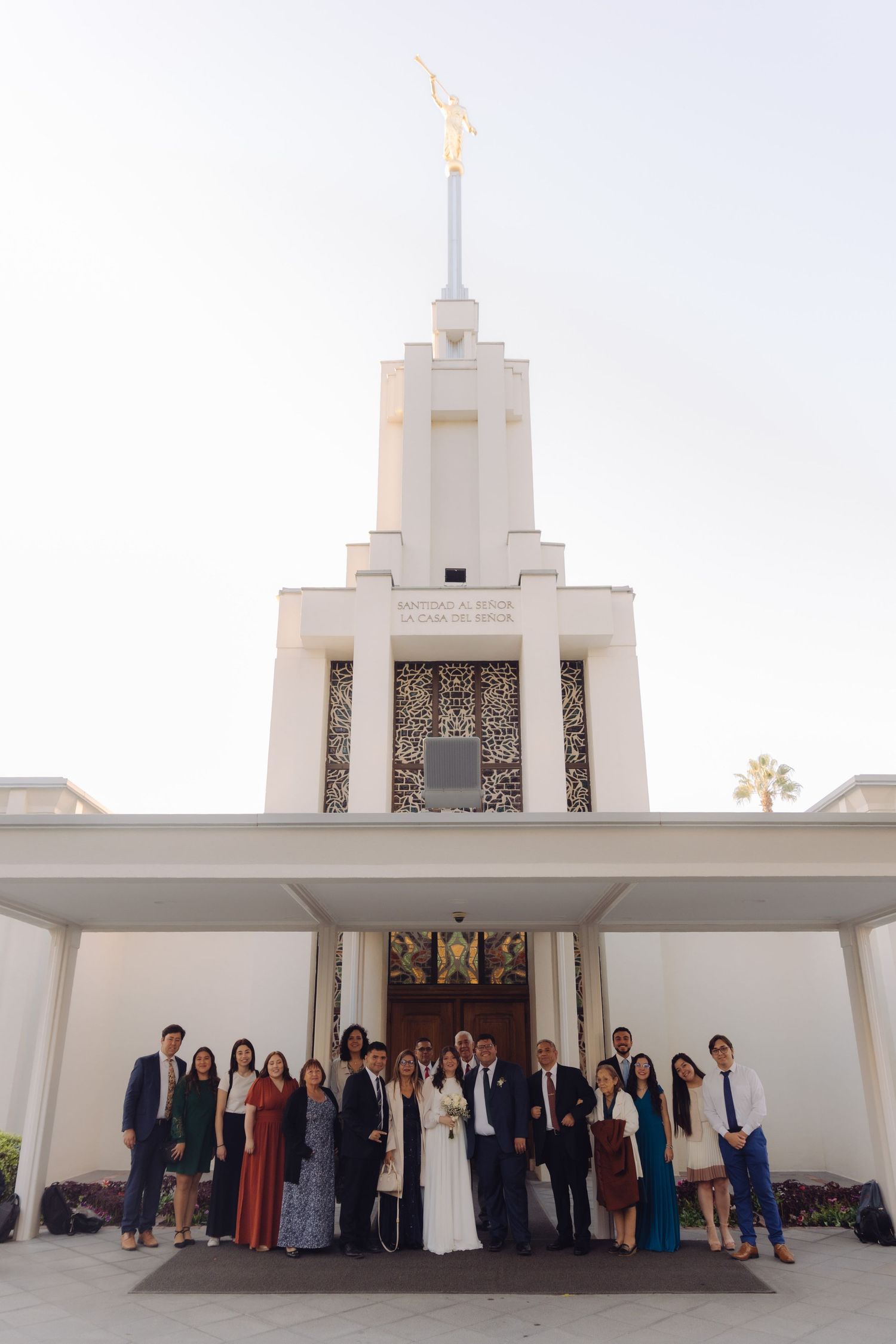 Large wedding group poses before a white Mormon temple with an angel Moroni statue on top.