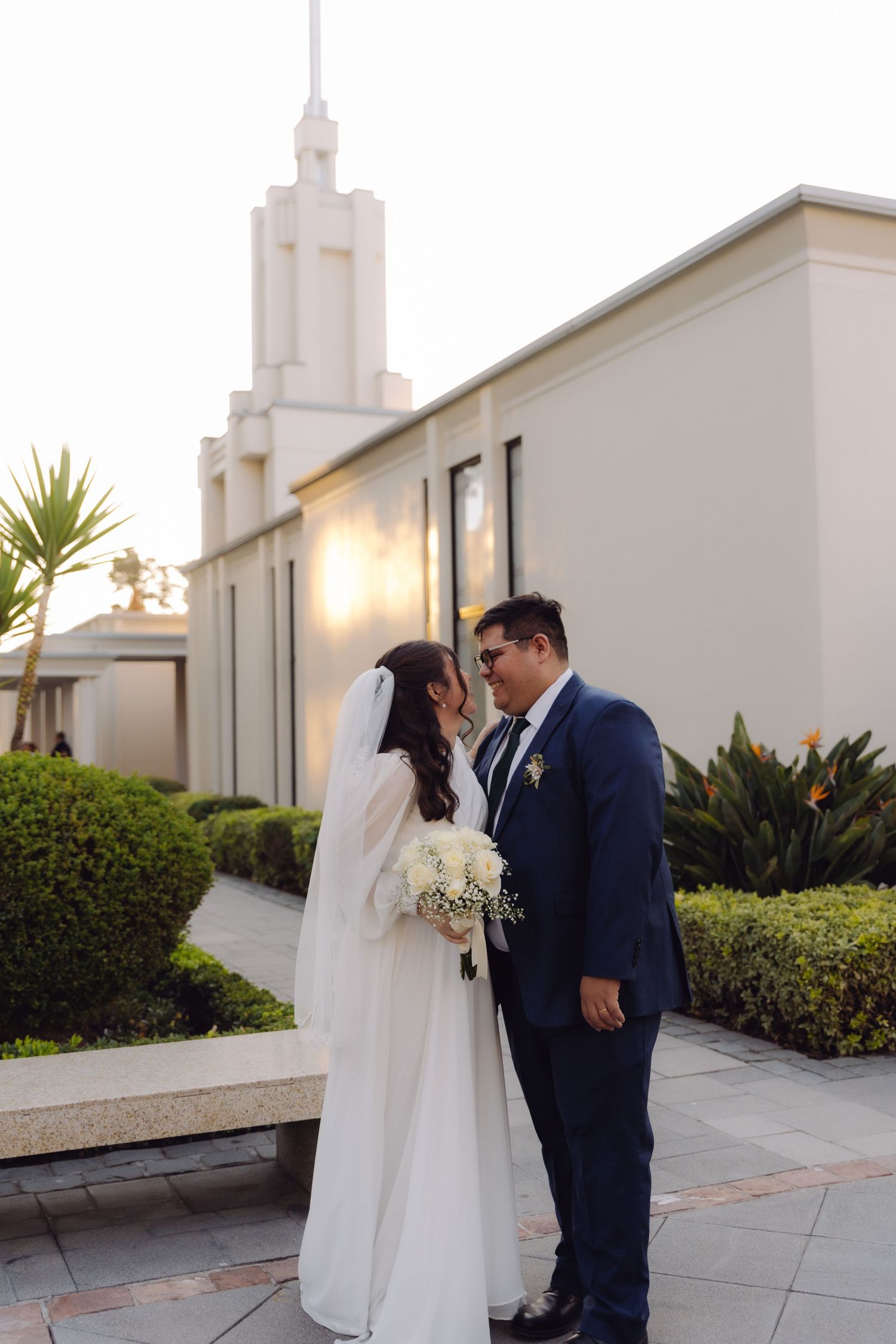 Newlywed couple gazes at each other holding white bouquet near a sunlit LDS temple exterior.