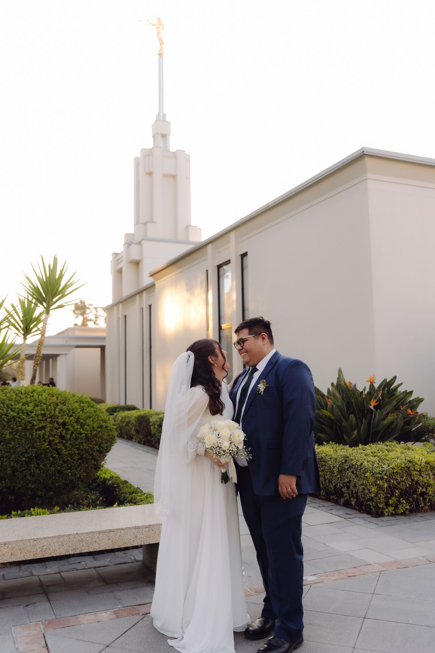 Bride and groom stand facing each other with white temple spire glowing in background at sunset.