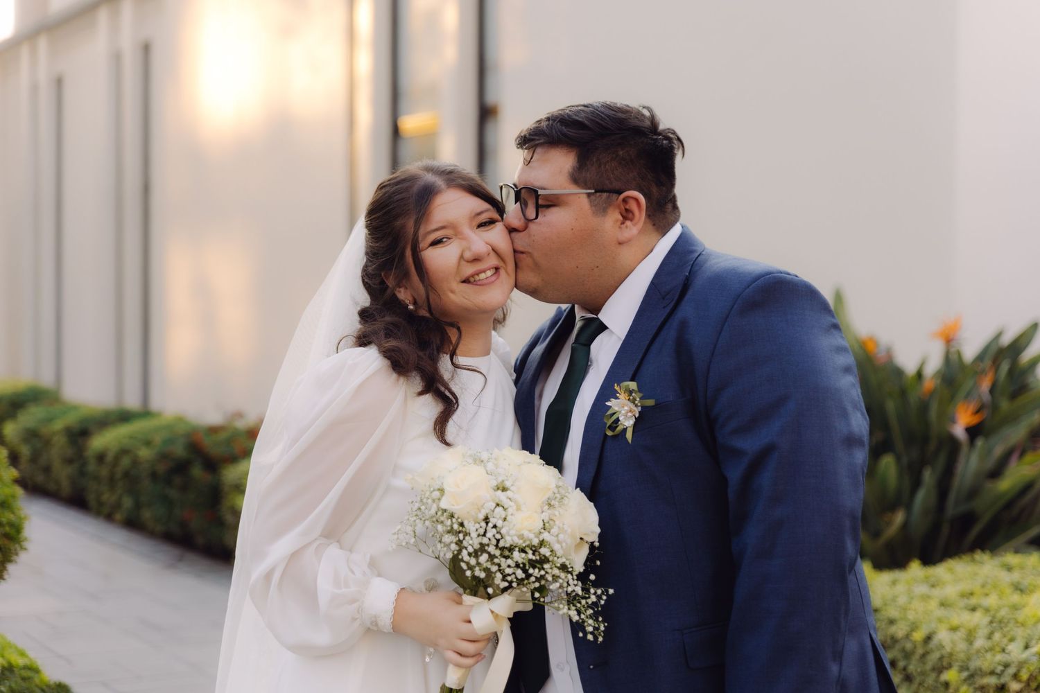 Groom kisses bride on the cheek as she smiles holding a white bouquet outside a temple.