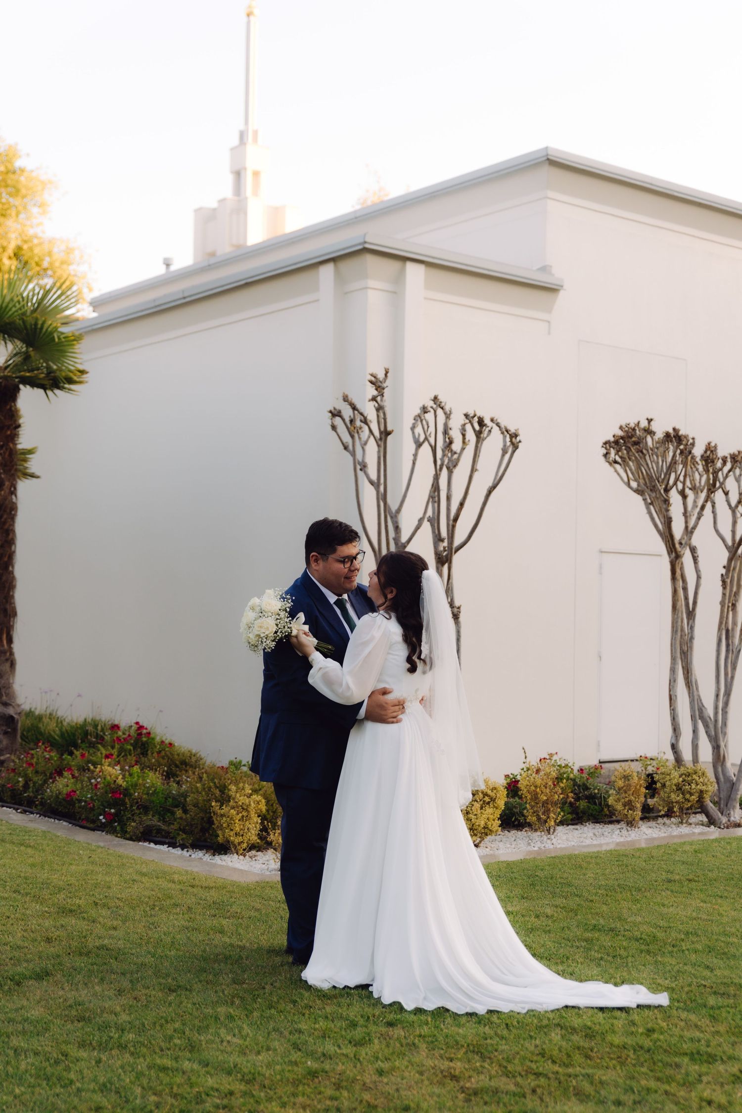 Bride and groom embrace on green lawn outside white LDS temple building with palm trees.